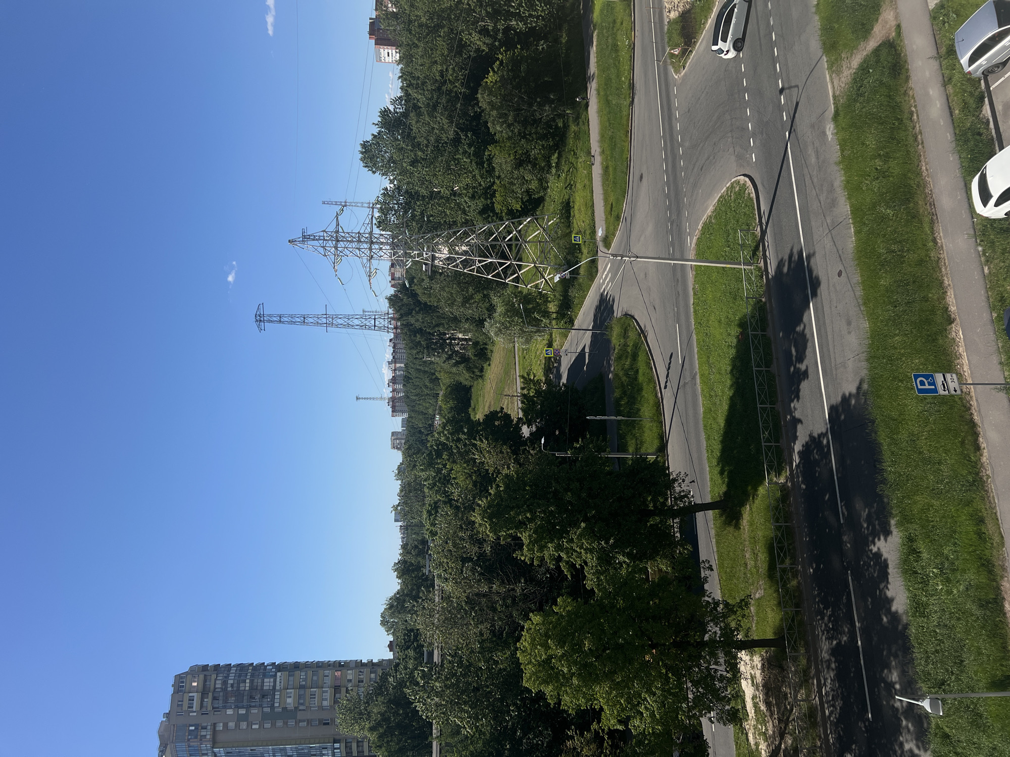 This image captures an urban landscape in Saint Petersburg, Russia, under a clear blue sky. In the background, several multi-story residential buildings are visible, interspersed with lush green trees. Dominating the mid-ground are two prominent electrical transmission towers, one significantly larger than the other. A winding road system with white lane markings cuts through the scene, with a white car navigating a curve. Green grass verges line the roads, and a pedestrian sidewalk is visible in the foreground. A parking sign with a "P" symbol and an arrow indicating a distance of 30 meters is also present. A lamppost stands near the intersection. The overall impression is of a modern, developed urban area surrounded by significant green spaces.