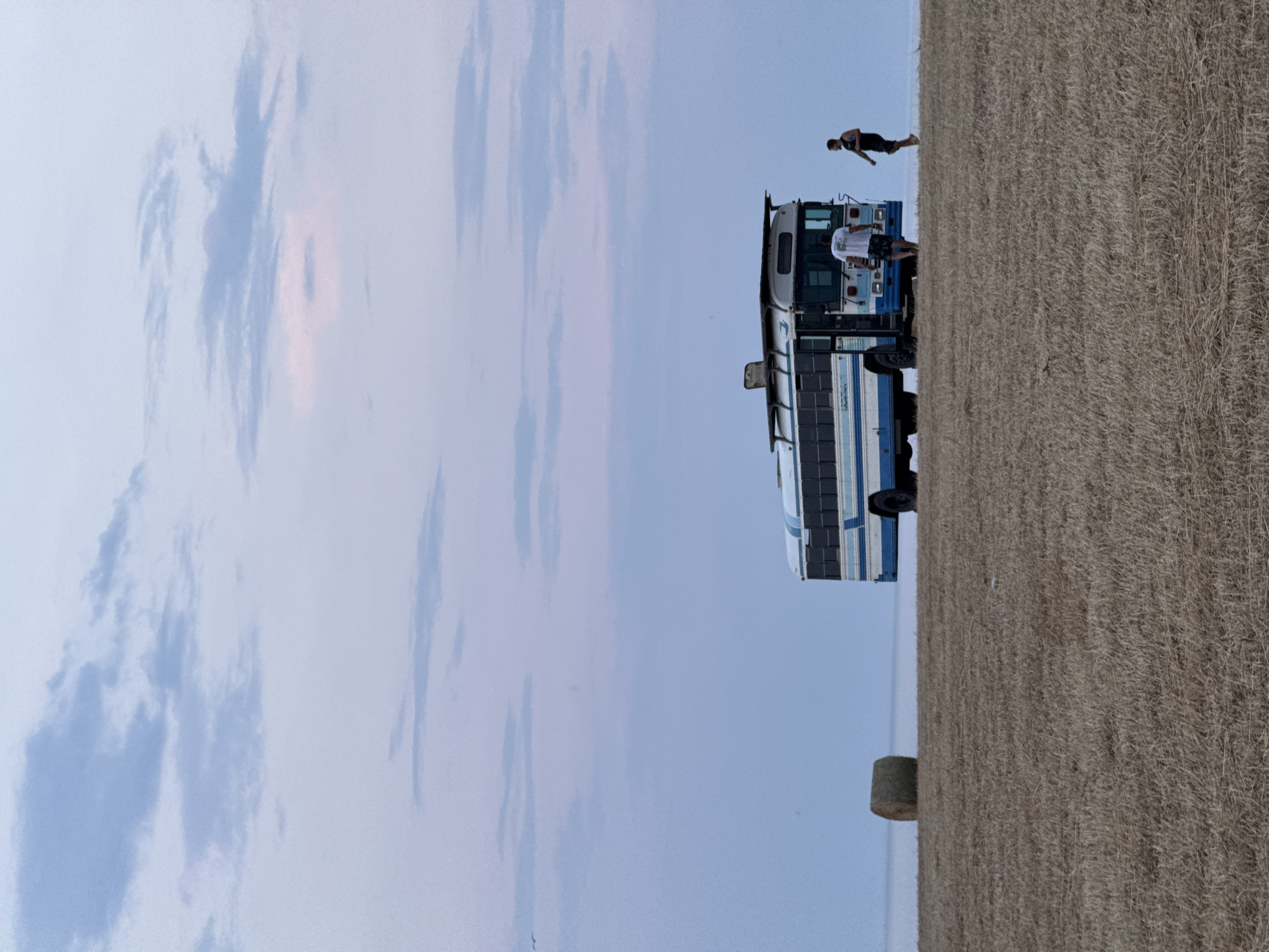 In a harvested field, under a pale sky with wispy clouds, a blue and white school bus is parked. To the left of the bus, a round bale of hay sits in the field. Two individuals are near the bus; one is facing away from the camera, looking into the open doorway of the bus, while the other, facing the camera, walks away from the bus. The field appears dry and recently harvested. The horizon is barely visible, suggesting a wide, open landscape. The scene is set in Varvara, Bulgaria.