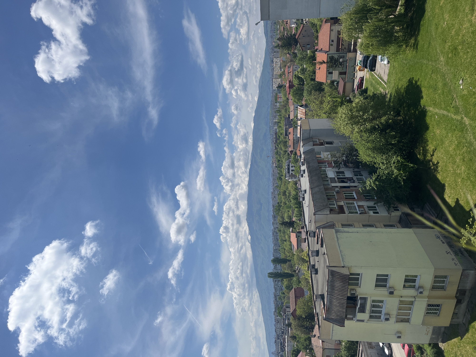 The image captures an elevated view of Sofia, Bulgaria, under a bright blue sky scattered with cumulus clouds. A mountain range forms a backdrop, partially obscured by cloud cover. The foreground consists of residential buildings with mixed architecture; some have orange tile roofs, while others are multi-story apartment blocks painted in light colors. Green trees are interspersed among the buildings. A small section of manicured grass is visible on the right. In the middle ground, various cars are parked in a residential area. Graffiti markings can be seen on the side of one of the buildings.