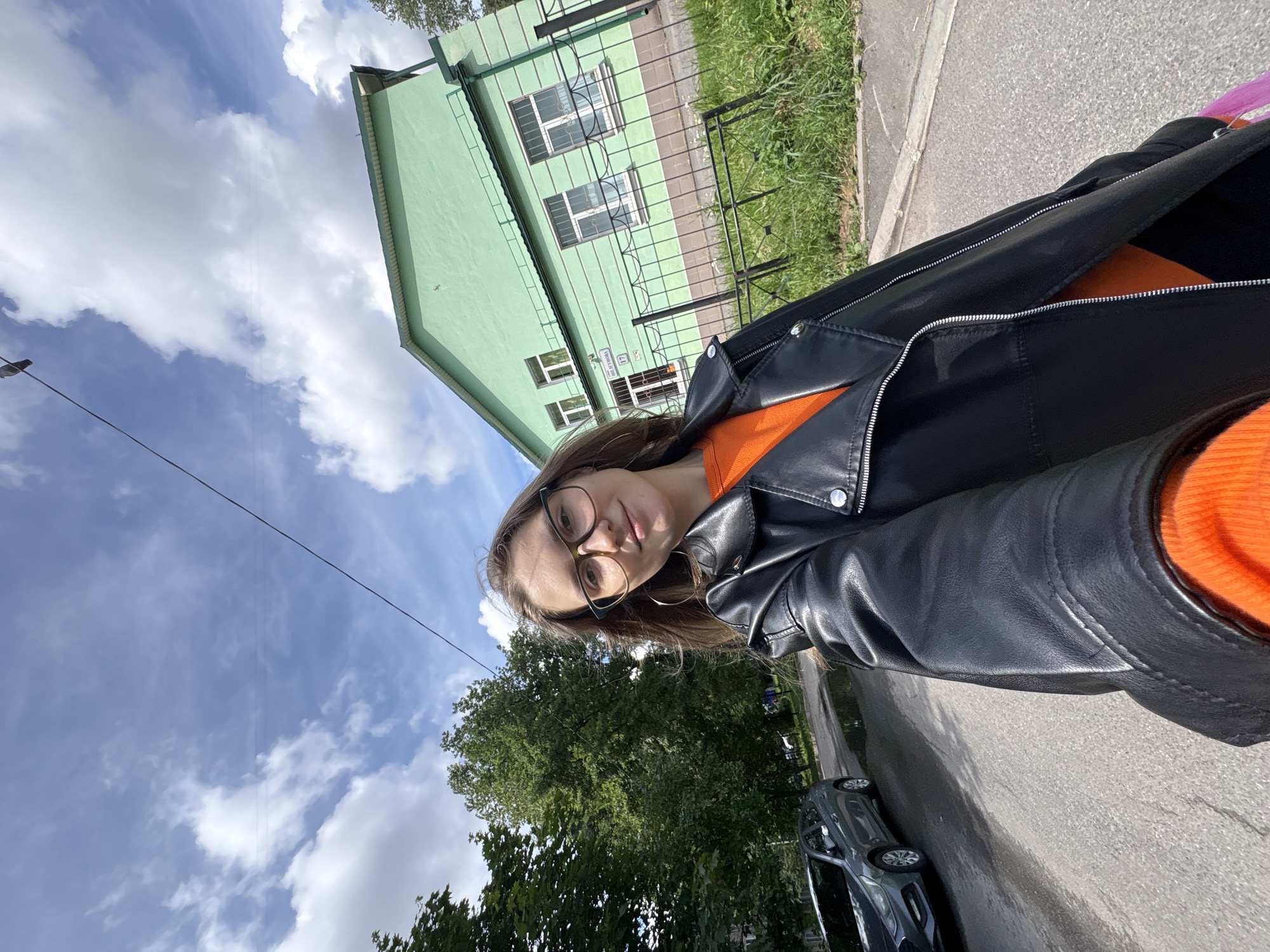 A young woman with brown hair and glasses is taking a selfie outdoors. She is wearing an orange top and a black leather jacket. In the background, there is a green building, trees, a street with a car, and a cloudy blue sky. A power line runs diagonally across the sky. The location is identified as Saint Petersburg, Russia, based on the street sign that reads "Шелкунова 17", which corresponds to Shelkunova Street, House 17 in Saint Petersburg.