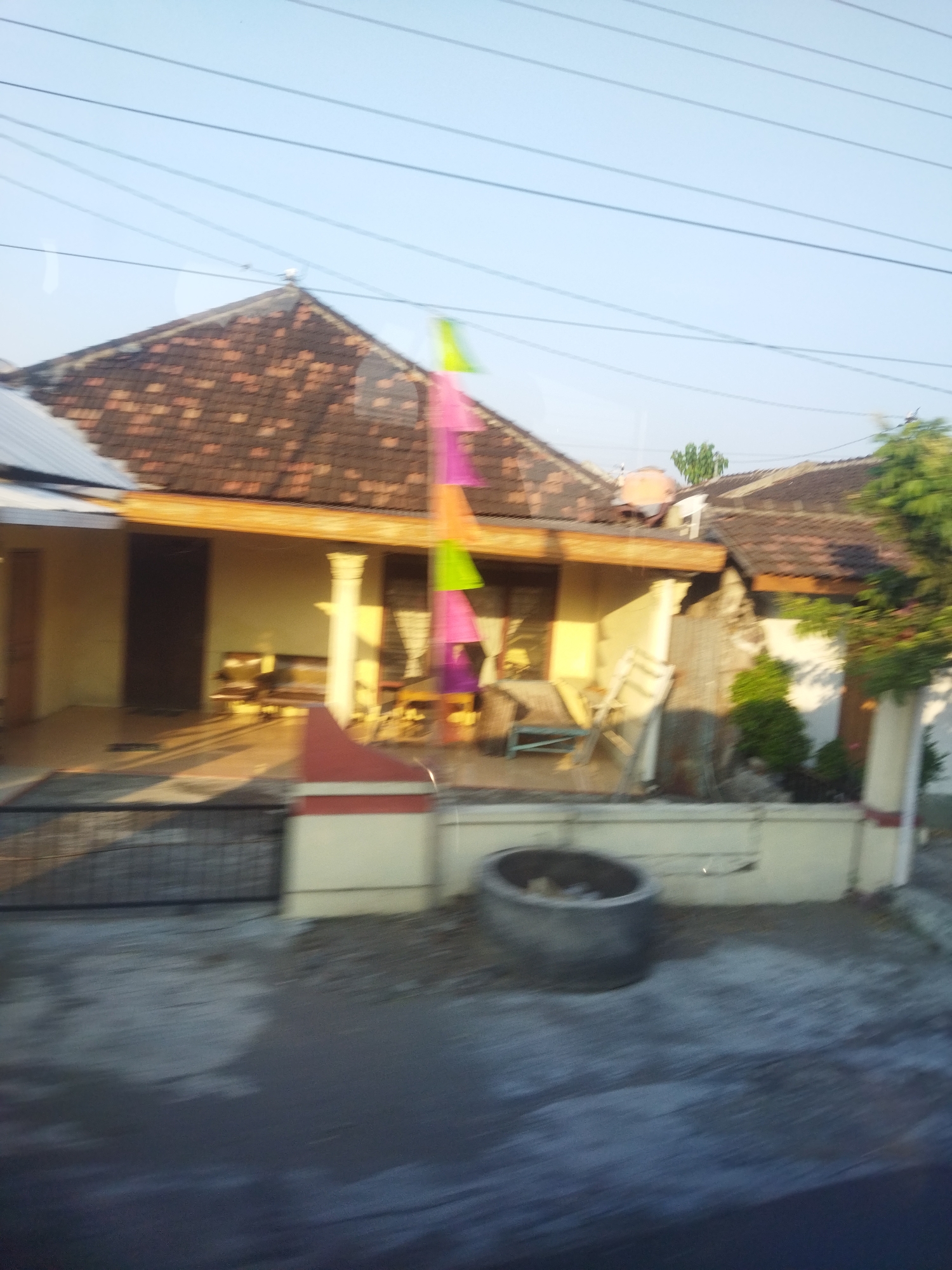 In the city of Selogiri, Indonesia, a blurred, horizontal image captures a residential scene. The main subject is a house with a tiled, pitched roof. In front of the house, a covered porch area features seating. Decorative triangular flags in pink, green, and purple hang on a pole. The ground in front of the house is paved, and a round, concrete structure, possibly a well or planter, is visible. A metal gate and a fence line the property. Power lines are strung across the sky. The environment suggests a clear day, with sunlight casting shadows.