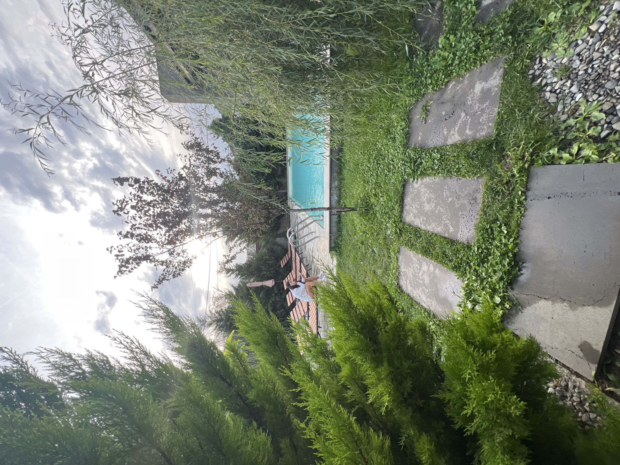 This image depicts an outdoor residential scene, likely a backyard, in Tsodoreti, Georgia. In the foreground, a pathway made of large, flat, grey stone slabs is surrounded by lush green grass and small pebbles. To the left, a vibrant green evergreen tree dominates the frame, its branches reaching towards the sky. To the right, a weeping willow tree with long, delicate branches hangs over the scene.
Beyond the pathway, a swimming pool with bright turquoise water is visible. Adjacent to the pool are several empty lounge chairs made of wood and fabric, some with beige cushions. In the middle ground, a young woman with brown hair is seated on one of the lounge chairs, wearing a white long-sleeved shirt and light-colored shorts. She appears to be looking down, possibly at her phone. Behind her, more lounge chairs are arranged, and a light-colored umbrella is partially visible. The background consists of trees and a modern-looking building with a dark grey facade. The sky above is partly cloudy with streaks of white and grey clouds, and the sun is casting a soft light. The overall atmosphere is serene and suggests a leisure setting.