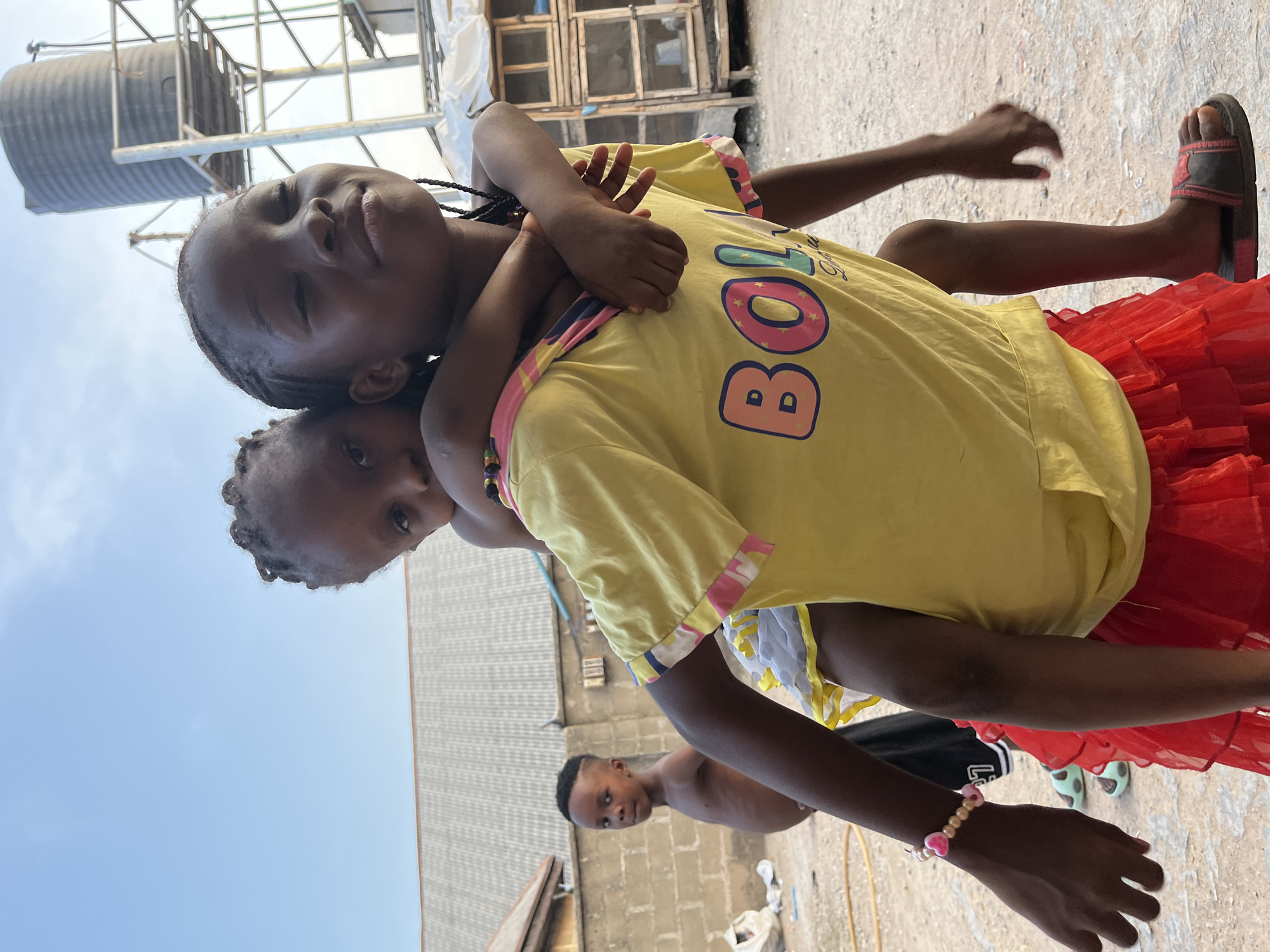 Two young girls are engaged in a piggyback ride in an outdoor environment in Gwagwalada, Nigeria. The girl giving the piggyback ride wears a yellow t-shirt with 'BOL' printed on it, a red tutu, and dark sandals. The girl on the back wears a white top. Another young boy, bare-chested, is partially visible in the background. The location features a water tank, surrounding buildings, and visible sky, indicating an urban or semi-urban setting.