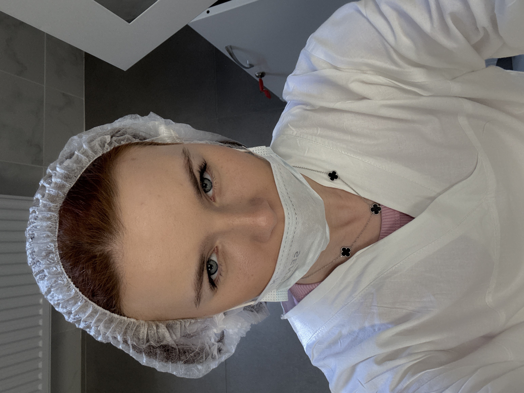 A woman wearing a white bouffant cap and a white medical mask is wearing a white medical gown, and appears to be in a medical or laboratory setting. She has light skin, light eyes, and brown hair. She wears a necklace with black quatrefoil pendants. The background includes a grey wall and light grey cabinets, which have a silver handle. The scene may be in Saint Petersburg, Russia.