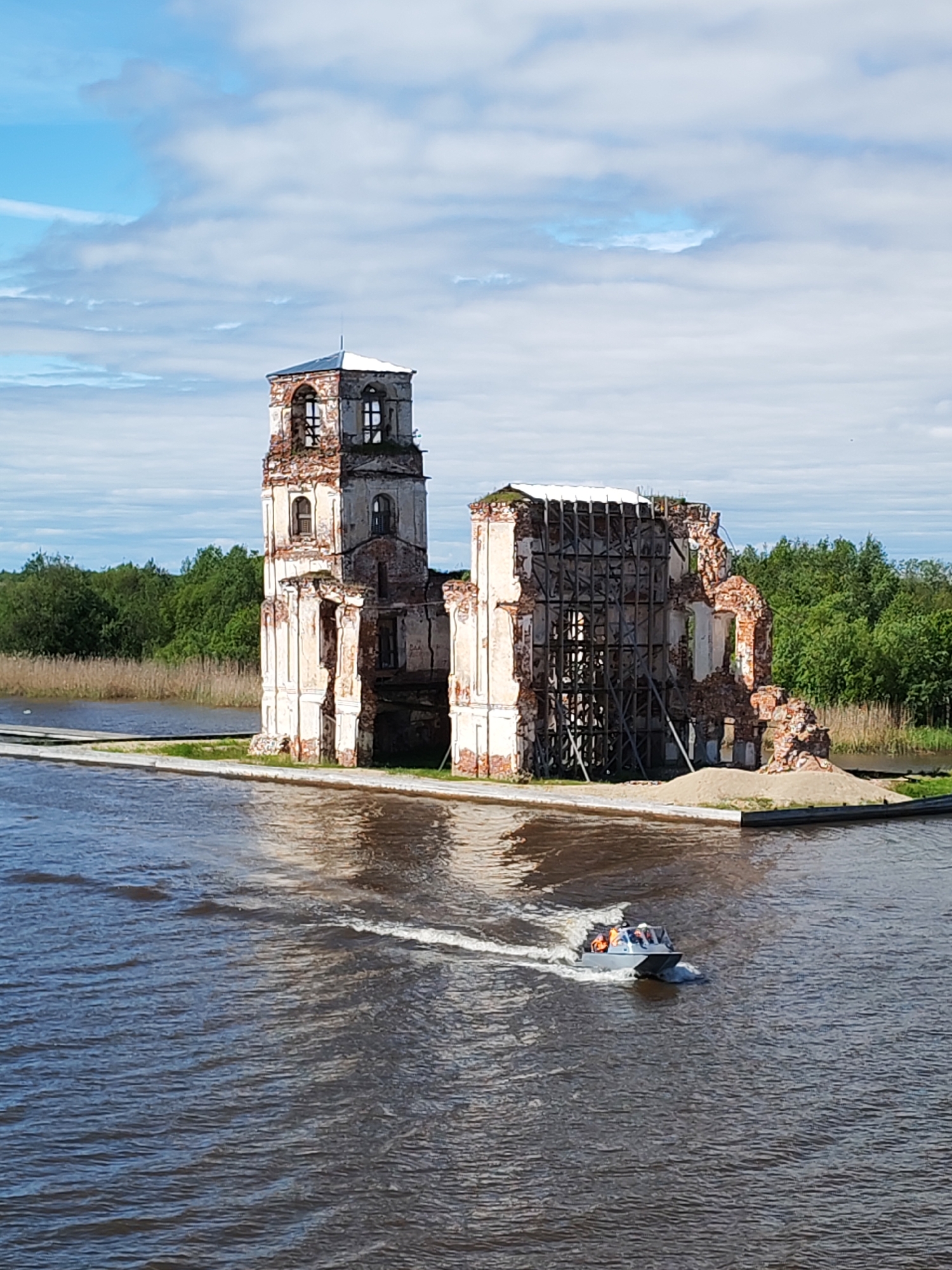 The image depicts the ruins of a church in Krokhino, Russia. The church, located on a small island in a body of water, appears dilapidated with significant portions of its structure missing or damaged. Wooden scaffolding supports one section of the remaining structure. A motorboat with several passengers is seen on the water near the church. The background includes a shoreline with trees and vegetation. The sky is blue with scattered clouds.