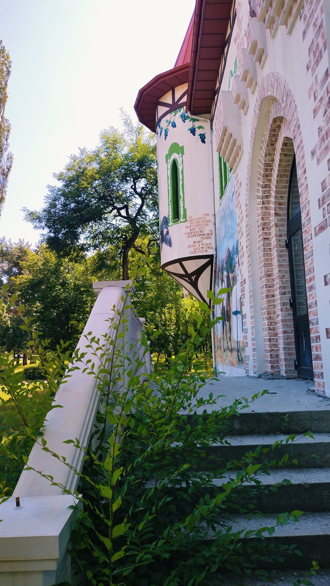The image depicts the exterior of a whimsical building with a curved, turret-like section adorned with painted green vines and blue grapes. This structure is made of light-colored stucco, accented with faux brick patterns and arched windows. A tall, dark-colored arched doorway is visible on the right side. The building's roof is a deep red. In the foreground, a white stone balustrade follows a set of concrete stairs leading upwards. Lush green foliage, including bushes and branches with small leaves, grows around the balustrade and stairs. The background features a dense canopy of green trees and a clear blue sky. The scene appears to be a park or garden area, likely in the city of Horishni Plavni, Ukraine, given the context. There are no people visible in the image.