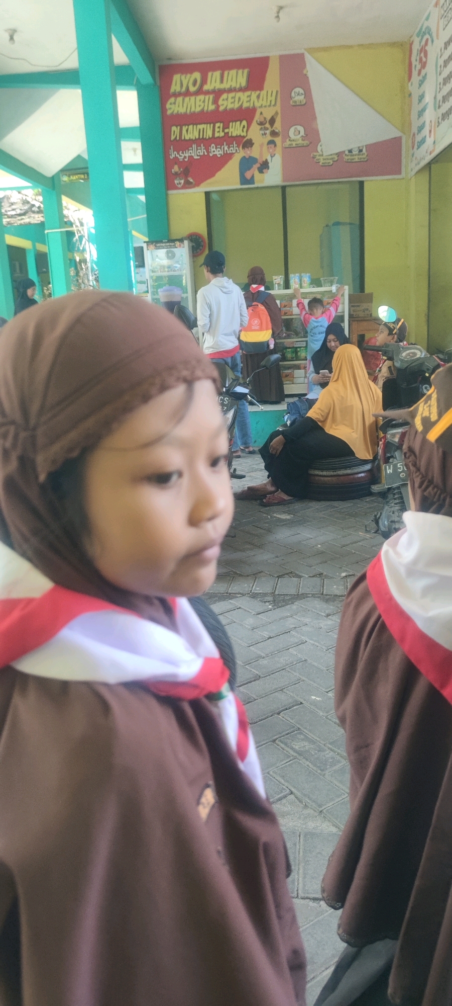In the city of Banjarsari, Indonesia, a close-up shot focuses on the side profile of a young girl wearing a brown headscarf and a brown scout uniform with a red and white sash. She is looking to her right, with her face partially obscured by the shallow depth of field. Behind her, other individuals are visible. A person in a white hoodie and a black baseball cap stands facing away. To their right, a person in a brown hijab and a bright orange and yellow backpack is also seen from behind. Further in the background, a woman in a yellow hijab is seated on stacked tires, holding a smartphone. Other individuals and parked motorcycles are present in the scene, suggesting a public outdoor area, possibly a school or community gathering. A banner with Indonesian text and cartoon illustrations is displayed on the wall behind the people, promoting shopping while donating. The ground is paved with interlocking bricks.