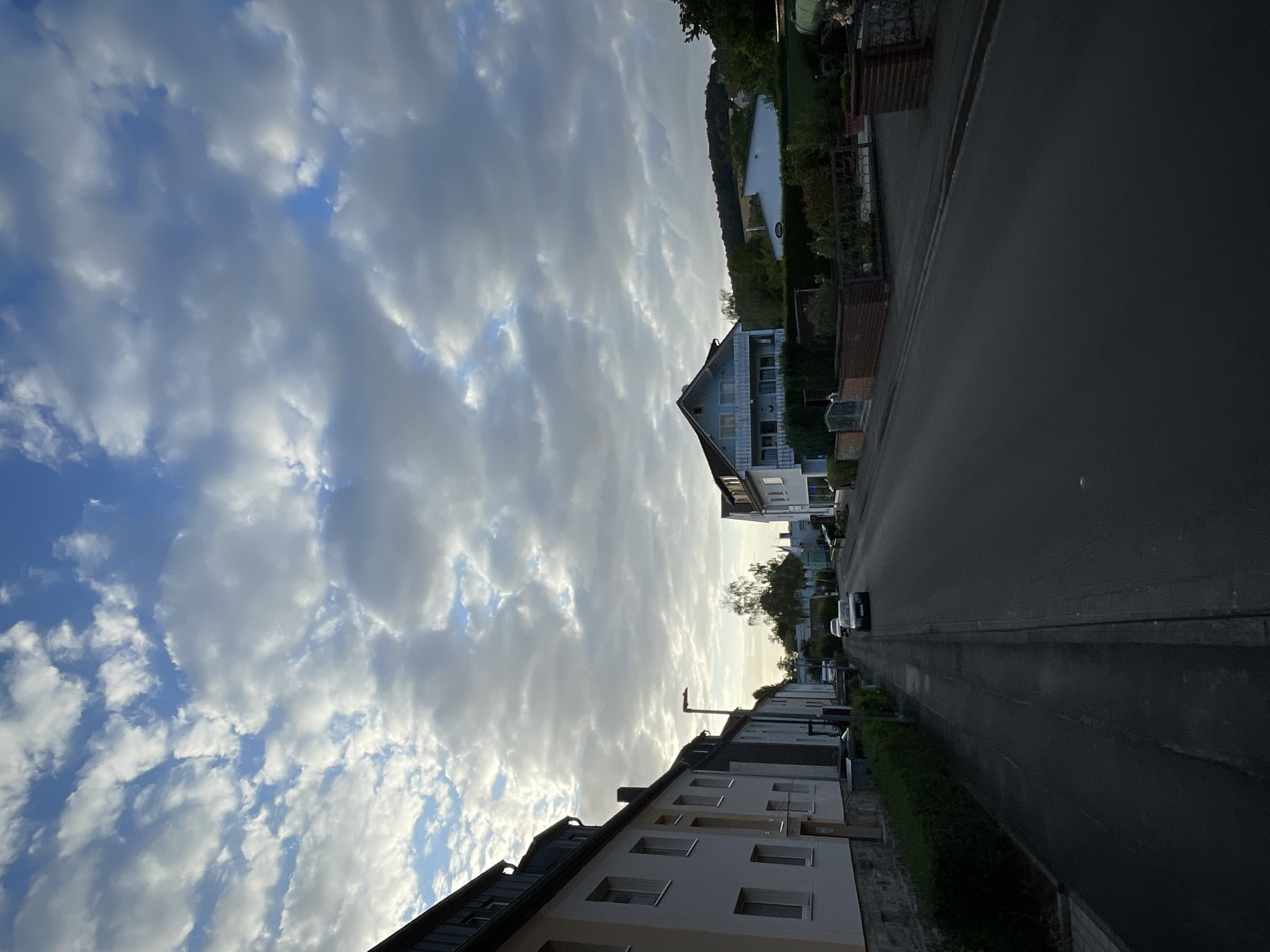 The image depicts a street scene in Neukirchen, Germany, under a sky filled with scattered clouds. On the left side of the street, there are several multi-story buildings with light-colored facades and dark-pitched roofs. A sidewalk runs alongside these buildings, bordered by a neat green hedge. Further down the street, a lamppost is visible.
The street itself is paved and appears to be a residential area. Several cars are present: a white car is parked, and a dark-colored car is driving away from the camera. On the right side of the street, more buildings are visible, including a light blue house with multiple windows and balconies. Behind the houses and buildings on the right, a gently sloping, tree-covered hill can be seen. In the distance, beyond the hill, a body of water is faintly visible. The overall atmosphere suggests a quiet suburban or residential neighborhood during daylight hours.