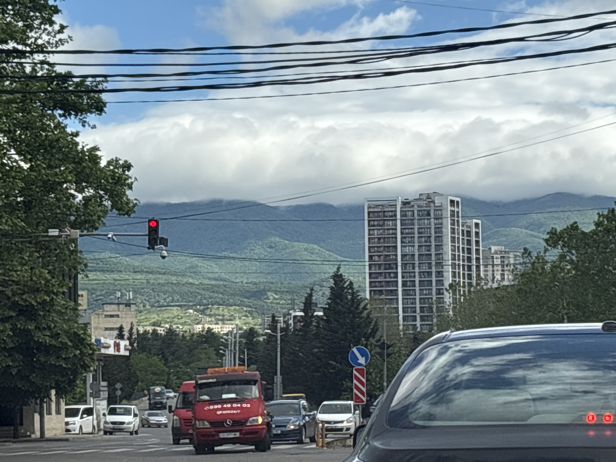 The image depicts a street scene in Tbilisi, Georgia, with a view of the city skyline and surrounding mountains under a cloudy sky. A red traffic light is visible on the left. Multiple vehicles are on the road, including a red tow truck with Georgian script and the phone number "596 49 04 03" displayed on its side. A tall, modern apartment building dominates the center-right background. Overhead, a network of power lines stretches across the sky. The image is taken from inside a vehicle, with the back of the vehicle visible in the lower right corner. Trees are on the sides of the street.