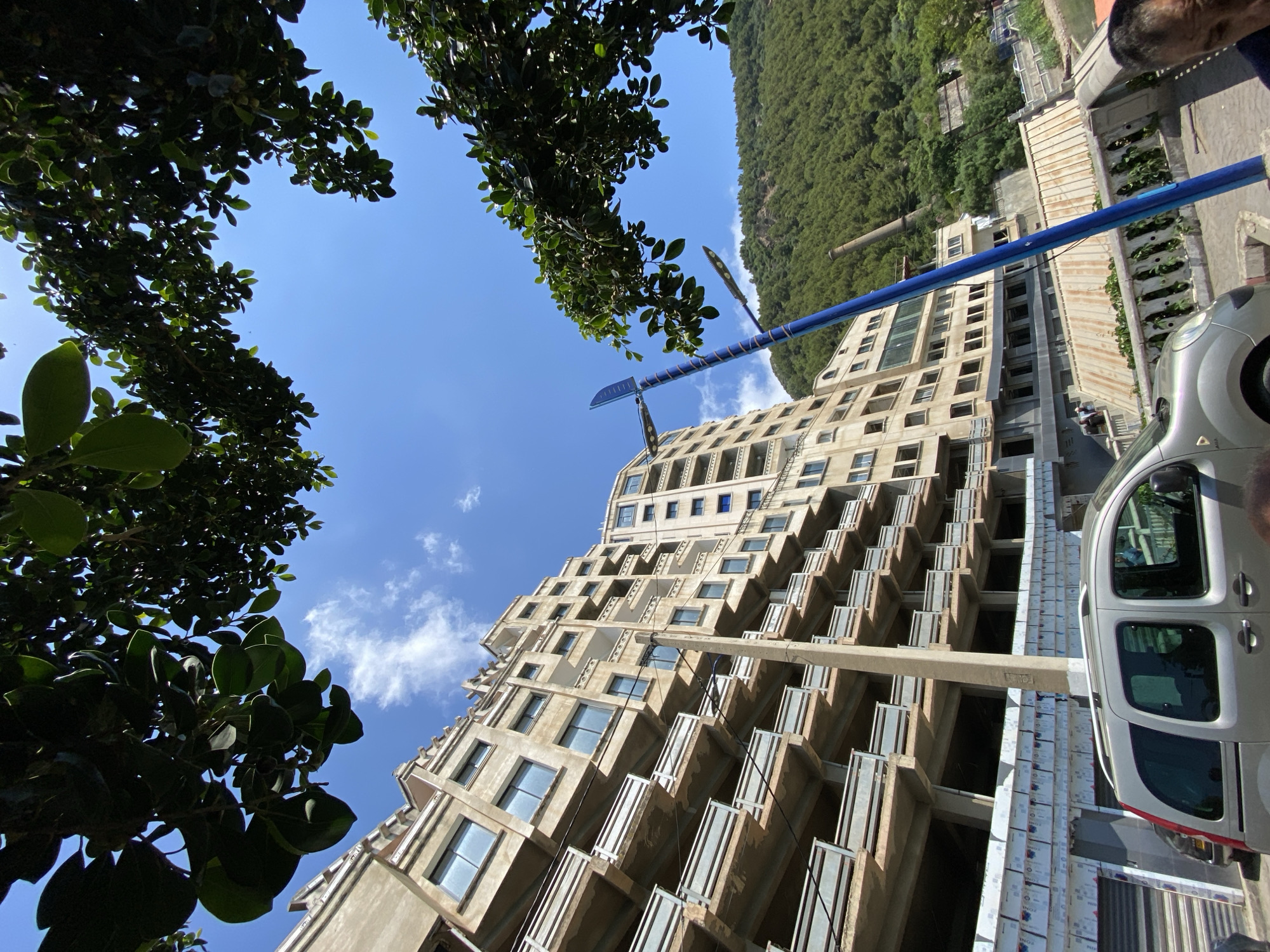 This media file presents a dynamic upward-looking view of a multi-story building, framed by lush green foliage and set against a bright blue sky. The image is captured from a low perspective, creating an impression of towering structures and expansive sky.
The primary subject is a tall, modern building with a distinct terraced or stepped design on its right side, featuring numerous rectangular windows and balconies. The building's facade is a light, possibly beige or concrete color, and appears to be in varying stages of completion, with some sections showing exposed concrete and others with finished windows.
In the foreground, dense green trees and foliage frame the top and left edges of the image, obscuring parts of the sky and adding a natural element to the urban scene. Above the building, a clear blue sky is visible, dotted with a few scattered white clouds. To the right of the building, a thickly forested, green hillside rises, suggesting a location where urban development meets natural landscape.
Various man-made objects populate the scene. A prominent, vibrant blue pole, possibly part of a decorative street light or art installation, extends diagonally across the mid-ground, featuring intricate, serrated-edge embellishments. Partially visible on the far right is the front section of a silver or grey car, possibly a van, parked near the building. Near the car and the base of the building, construction materials wrapped in white plastic are visible, displaying repeated text that includes "ZONE CONTROL".
The bright sunlight and clear sky indicate that the photo was taken during the daytime, likely mid-morning or early afternoon, under pleasant weather conditions. The combination of a modern building, natural greenery, and ongoing construction suggests an area undergoing development or revitalization. The location cues, such as the architecture and forested hill, align with the provided context of Bejaia, Algeria.