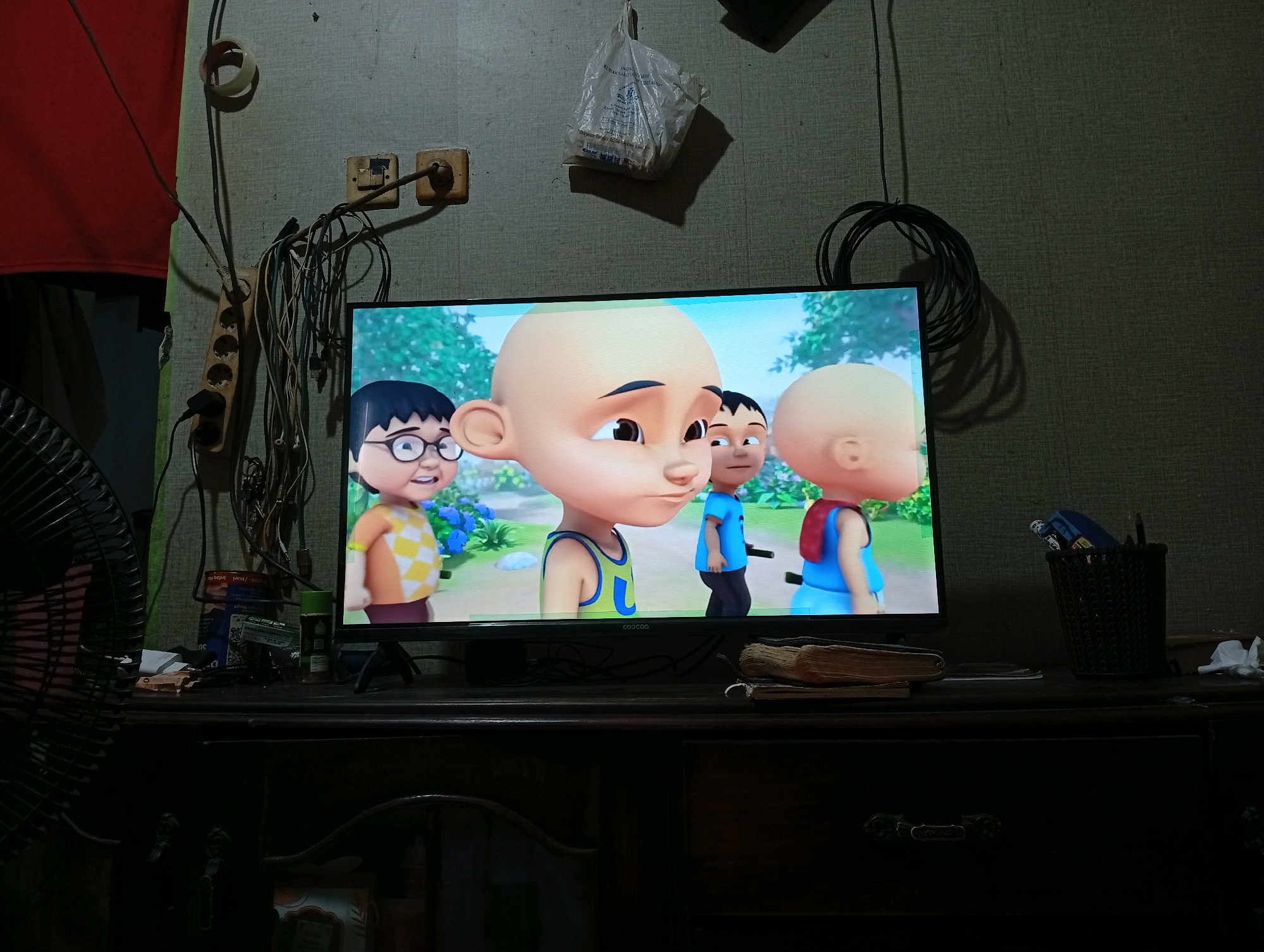 The image depicts an indoor scene with a television displaying an animated cartoon. The television is placed on a dark wooden cabinet. To the left of the television, a fan is partially visible. Above and behind the television, the wall is textured and a collection of electrical wires and sockets are visible, along with a plastic bag hanging from the wall. To the right of the television, a pencil holder filled with pens and pencils is on the cabinet, alongside a crumpled white object. The cartoon on the television features several animated characters with large heads and simplified features. The overall impression is of a domestic setting, possibly a living room, with a slightly cluttered and utilitarian feel, given the visible electrical setup. The context of "Caringin, Indonesia" suggests this scene is located in a residential area within that city.