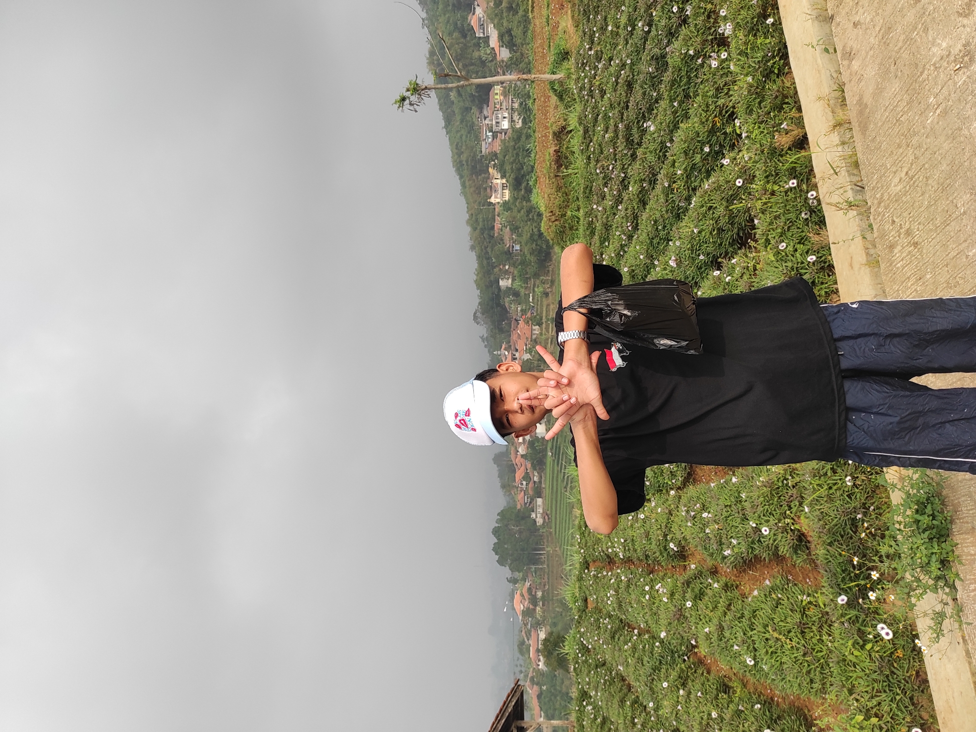 A young person wearing a white baseball cap, black t-shirt with a red and white logo, and dark blue athletic shorts stands in front of a field of small white flowers. They are holding their hands up, fingers extended, in front of their chest, and a black plastic bag is held in their right hand. Behind the field, there is a village with houses nestled on a hillside, and the sky is overcast and grey. The scene is likely in a rural area outside of Sumedang, Indonesia, given the architecture and vegetation.