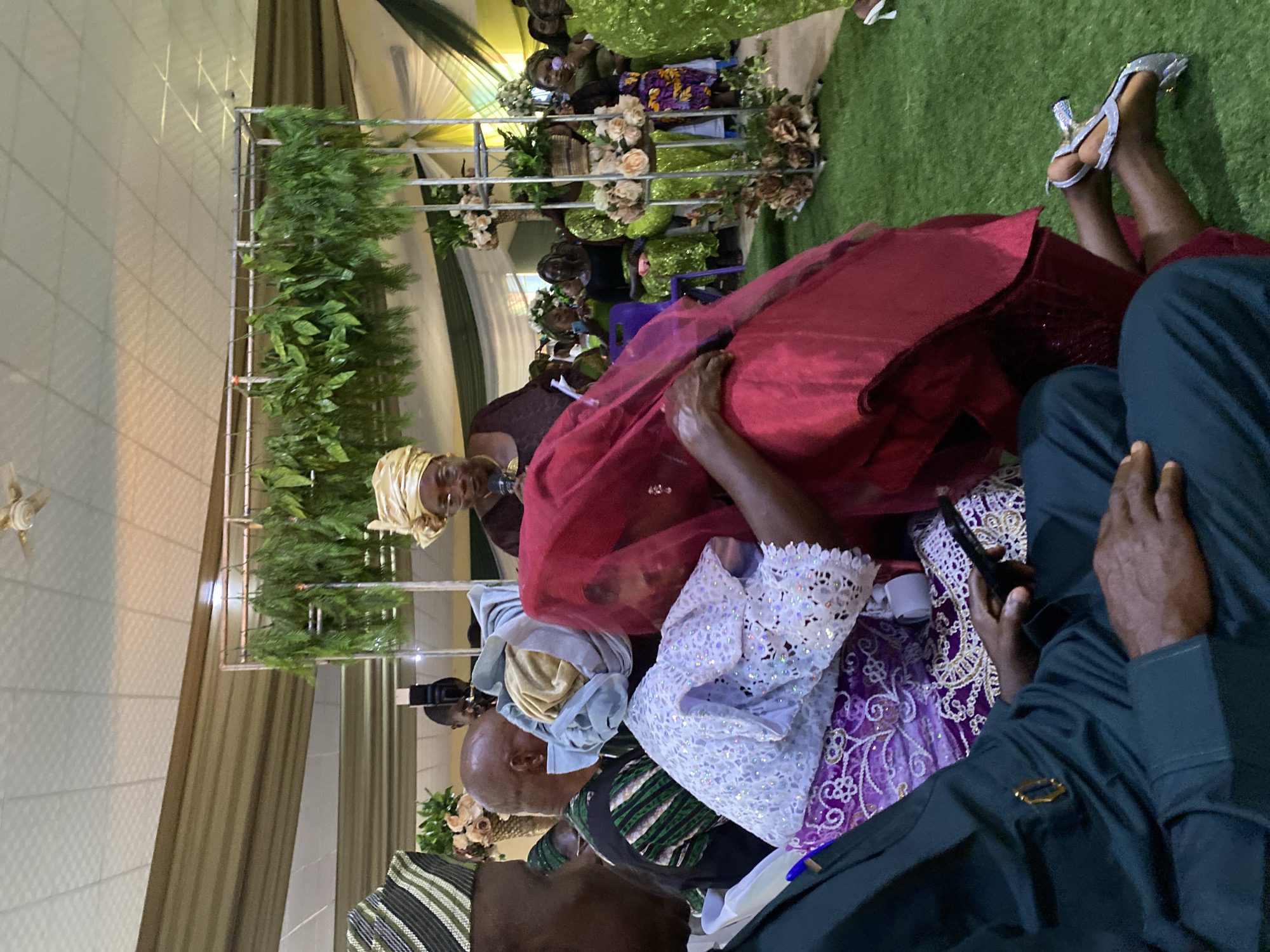 The image shows an indoor gathering in what appears to be Kuru Karama, Nigeria. Multiple people are visible, some wearing traditional attire including headwraps and colorful patterned clothing. A woman in a red covering is leaning on or being supported by another person. Others are seated. There is a decorated structure with greenery and flowers in the background, possibly an altar or stage. Someone is holding a mobile phone. The environment includes artificial green turf and draped fabric.