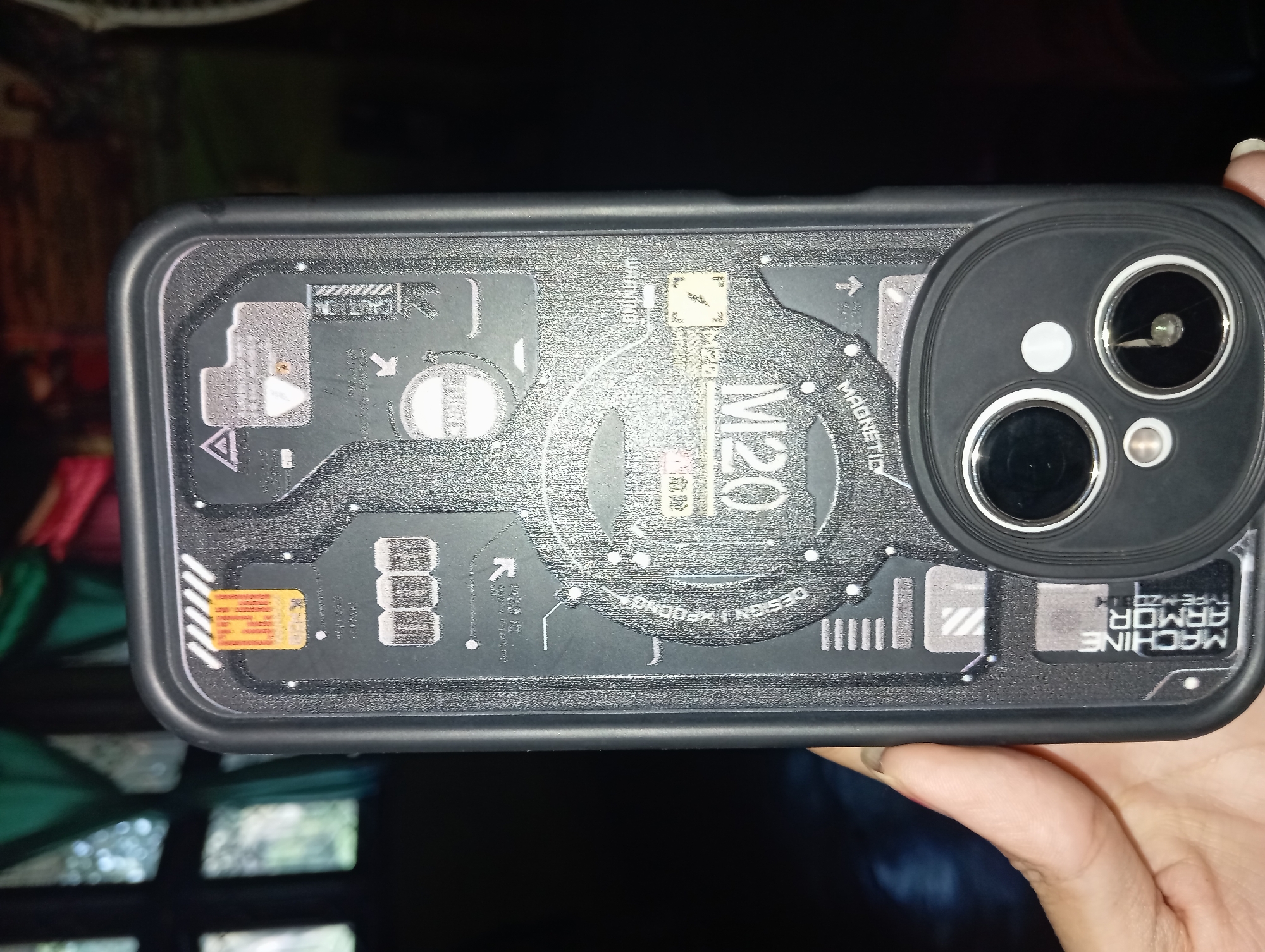 The image shows a person's hand holding a black phone case with a "Machine Armor" design. The case has various graphic elements resembling circuitry and warnings, with text like "M120" and "MAGNETIC" visible. The background is dark and out of focus, with some hints of green curtains and blurry shapes, suggesting an indoor setting with some natural light filtering in. The location is stated to be in Rejosari, Indonesia.