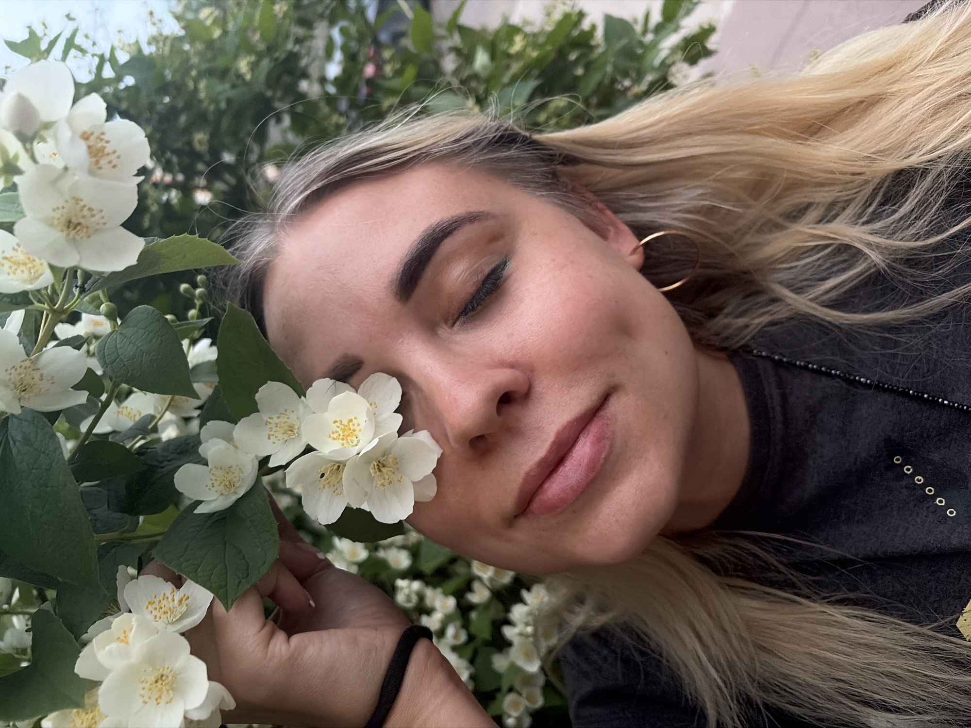 A close-up, medium shot captures a young woman with blonde hair positioned outdoors amidst white jasmine flowers. Her right eye is closed, with a few white jasmine blossoms obscuring it. Her left eye is open, looking towards the camera. She is wearing a dark grey t-shirt and a gold hoop earring on her visible left ear. Her hand is visible near the top left of the frame, gently holding some of the jasmine flowers. The background consists of lush green foliage and more white jasmine flowers, suggesting a garden or park setting. The image is taken in Chaikovsky, Russia.