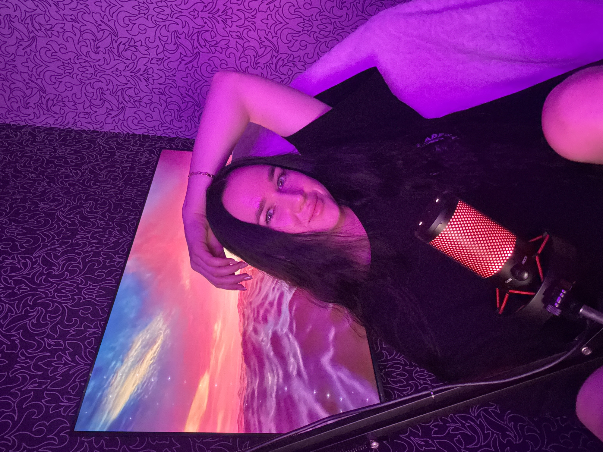 A person is lying on their side in a room with purple ambient lighting. The person has long dark hair and is wearing a black t-shirt. They are resting their head on their arm, which is bent at the elbow. Their eyes are open and they are looking towards the camera with a slight smile.
In front of the person is a large, flat monitor displaying a vibrant image of a colorful sky with clouds, rendered in shades of pink, orange, and blue. To the right of the person and the monitor is a black microphone with a red light, mounted on a stand.
The background consists of patterned wallpaper in shades of purple and darker purple, with a textured surface. The overall impression is that of a personal space, possibly a gaming or streaming setup, illuminated by mood lighting. The image conveys a relaxed and intimate atmosphere.
Visible text: On the microphone, the words "MODE" and "GAIN" are visible next to their respective dials. There is also faint, partially obscured text on the person's t-shirt.
The scene appears to be indoors, and while no specific time of day or weather is indicated, the purple lighting suggests it could be nighttime or a deliberately atmospheric setting. The location context is Novosibirsk, Russia.