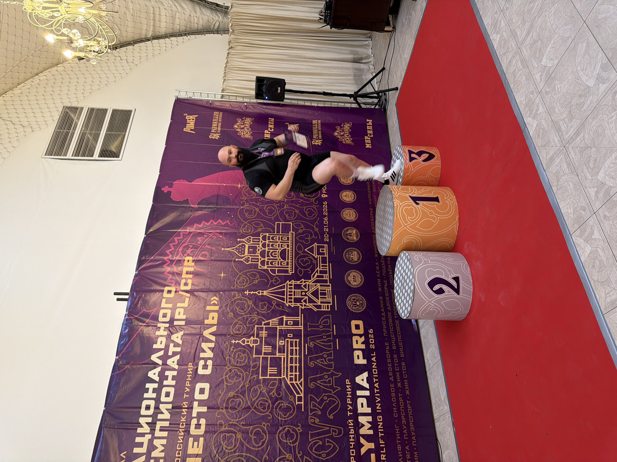 A muscular, bald man with a beard, wearing black athletic shorts and a black sleeveless top with a medal around his neck and holding a certificate, is captured mid-stride on a podium. He is on the second step, which is cylindrical and decorated with a pattern and the number "2". To his left is another podium step, also cylindrical, decorated with an orange pattern and the number "1". To his right is a third podium step, decorated with an orange pattern and the number "3". The podiums are on a red carpet that extends across the foreground. Behind the man is a large purple banner with gold ornate designs, depicting buildings that resemble Russian Orthodox churches, and text. The banner also includes logos for "POWER", "PAINKILLER HARDCORE DIVISION", and "DESPERADO". The text on the banner indicates the event is related to a "NATIONAL CHAMPIONSHIP IPL/SPR", a "RUSSIAN TOURNAMENT", and "OLYMPIA PRO - POWERLIFTING INVITATIONAL 2026". The backdrop suggests an indoor venue, possibly a hall or arena, with a high, arched ceiling visible at the top. To the right of the podiums, a black speaker on a tripod stand is visible, along with a small wooden cabinet. The man appears to be celebrating or posing, looking slightly upward and to his right. The overall scene depicts an awards or competition ceremony.
