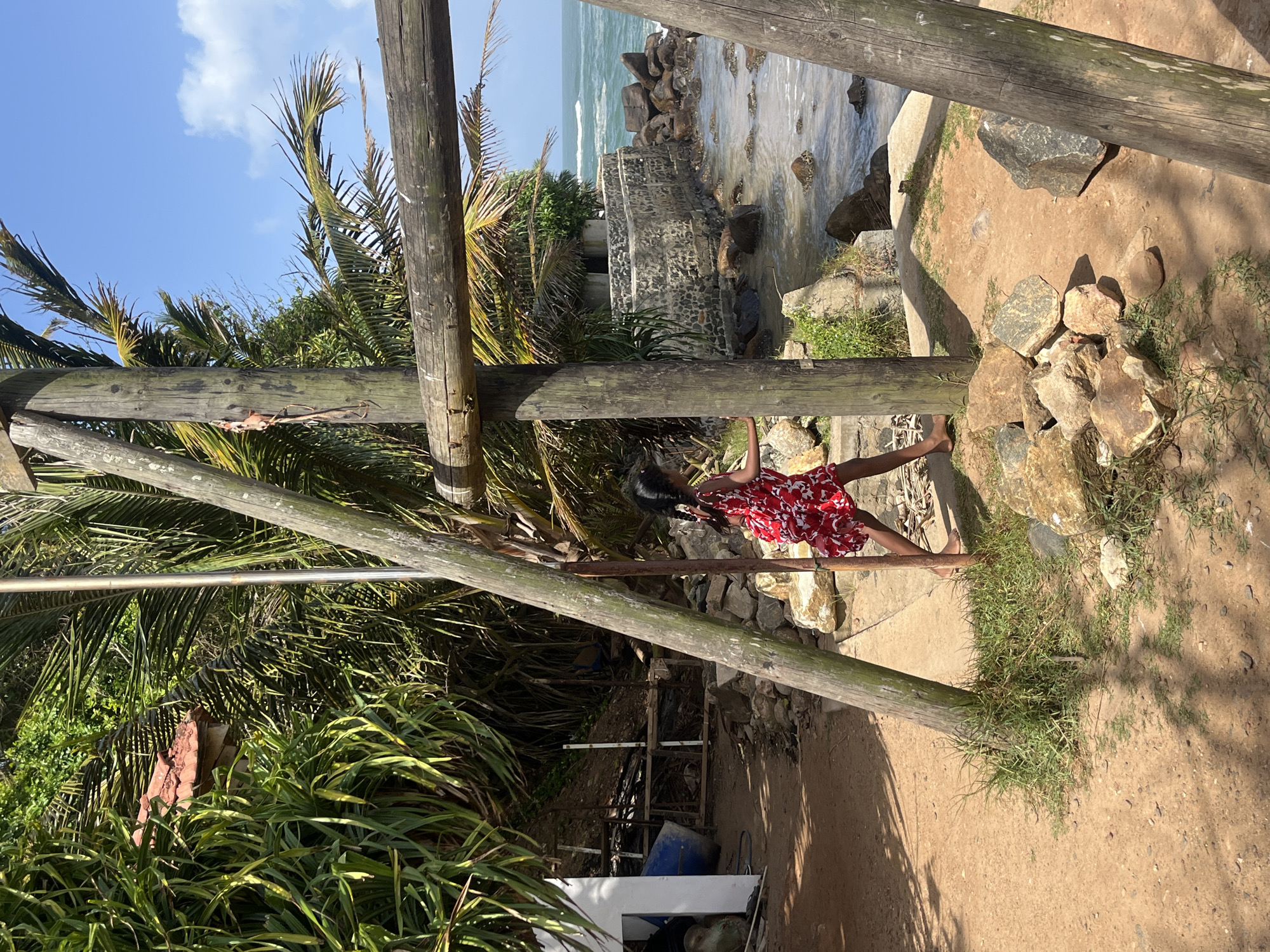A young girl in a red and white patterned dress walks to the right in an outdoor area with a sandy ground, rocks, and vegetation. She has dark hair in braids. In the background, there are palm trees, a structure made of stone and concrete, and the ocean with waves. Wooden posts and beams are present in the foreground, casting shadows on the ground. The scene appears to be a coastal area in Walgama, Sri Lanka.