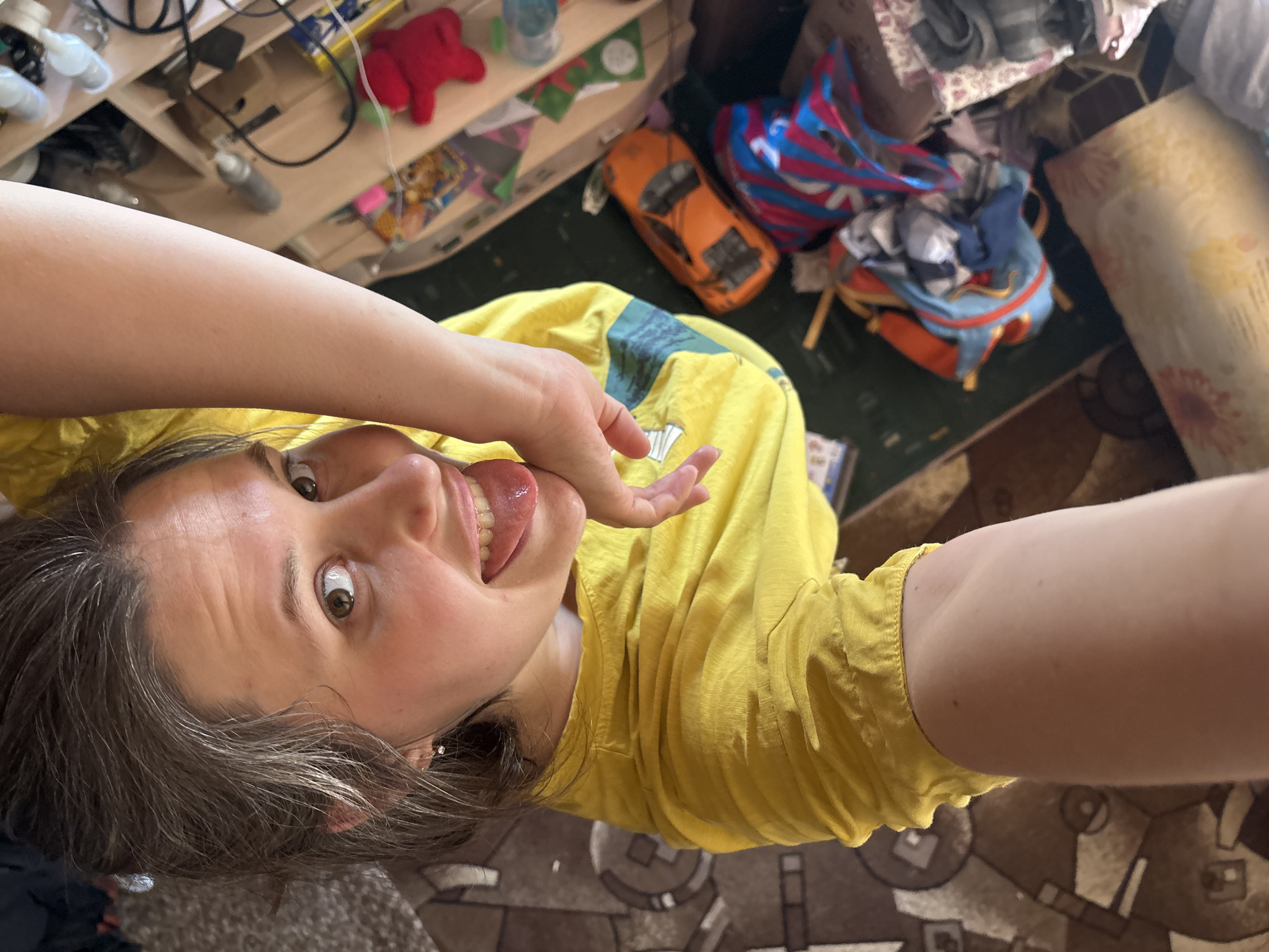 A high-angle, selfie-style photograph shows a fair-skinned woman with brown hair and eyes, wearing a yellow top, with her tongue partially extended. Her right hand is near her face. The background includes a cluttered room, with various objects like toys (an orange car and a red teddy bear), bags, and furniture visible. The location is possibly Nizhnyaya Chermoda, Russia, inferred from metadata, but there is no specific confirmation from the image itself. The scene appears indoors.