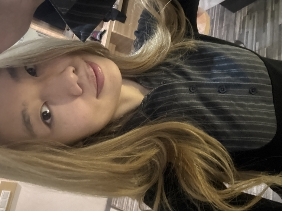 A selfie-style portrait of an East Asian female is captured indoors within Yuzhong District, China. The individual, positioned centrally, has light-toned skin, dark eyes, and long, wavy blonde hair parted to her right and falling over both shoulders. She exhibits a slight, closed-mouth smile while looking directly at the camera. She is dressed in a dark collared shirt featuring thin, light-colored vertical stripes, buttoned down the front, layered over or combined with a plain dark garment. Her right arm or hand is partially visible in the upper right quadrant of the frame, consistent with holding a device for self-photography. The background is an interior space with artificial lighting. A light-colored rectangular wall fixture with two square buttons is visible in the upper left. To the right, a blurred wooden table or furniture piece is present, on which indistinct dark items, possibly clothing, are placed. A patterned floor with geometric elements is faintly discernible in the lower background. The scene depicts a solitary individual in an indoor setting.