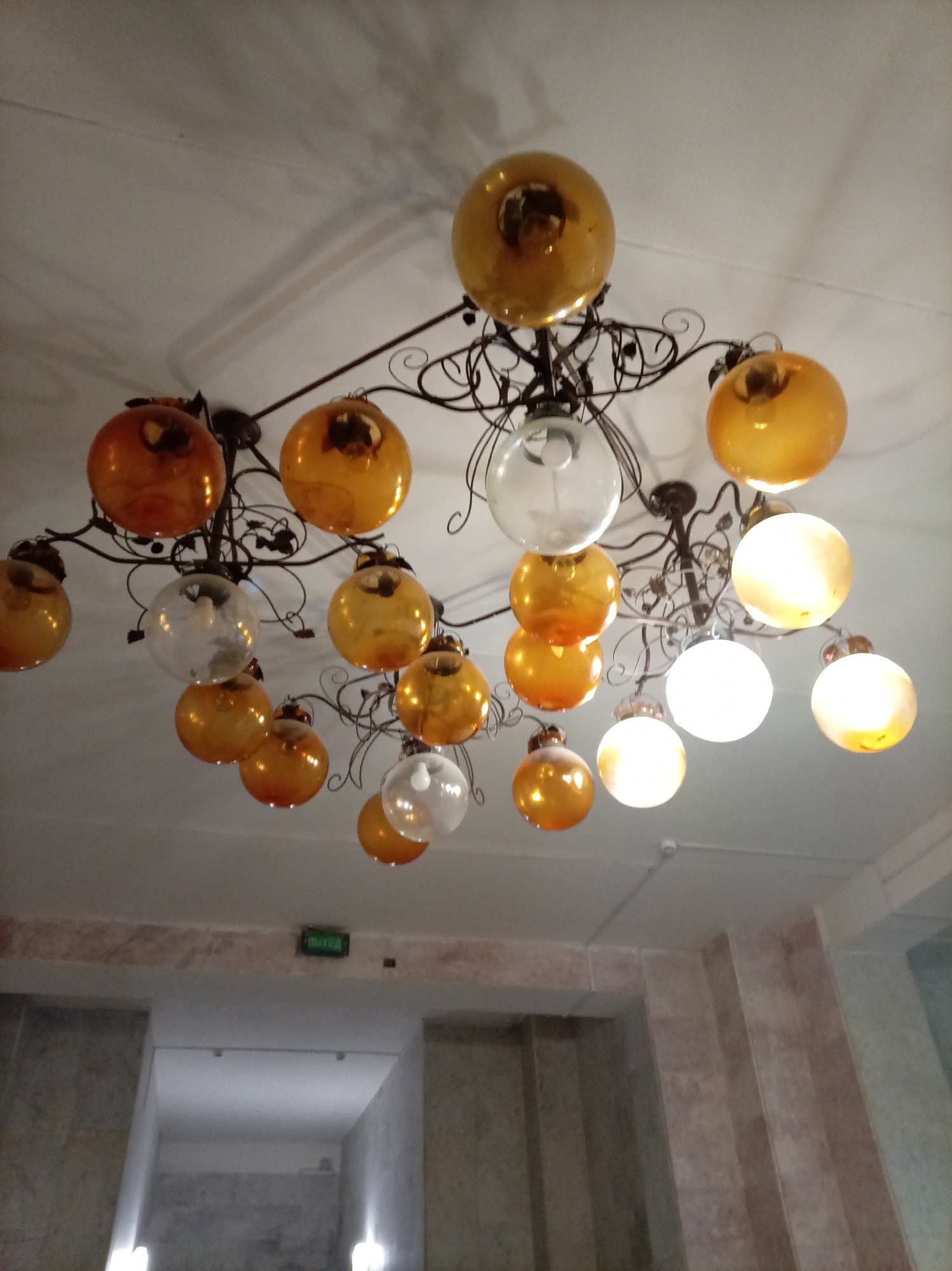 A chandelier with many amber-colored glass globes hangs from a white ceiling. The chandelier has a dark, ornate metal frame. Some of the globes are lit, while others are not. Below the chandelier, an illuminated green sign that reads "ВЫХОД" (EXIT) is mounted on a light-colored wall. The background shows a part of a building's interior, possibly a hallway or staircase, with textured walls and lights. The scene is likely indoors in Zheleznovodsk, Russia.