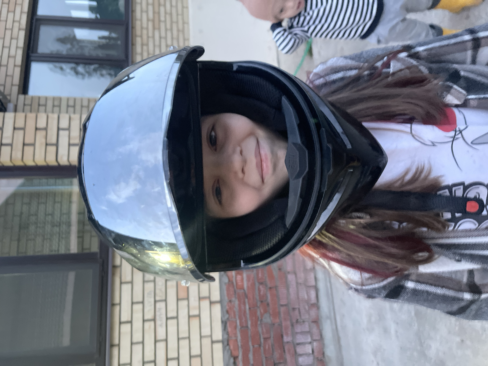 A girl with long brown hair, wearing a white t-shirt with a print, and a gray plaid shirt, is wearing a black motorcycle helmet. The helmet visor is up, and the girl is looking at the camera. A small child wearing a striped shirt can be seen in the background. The background includes a brick building with windows and a concrete surface. Based on available information, the location is in the city of Chaikovsky, Russia.
