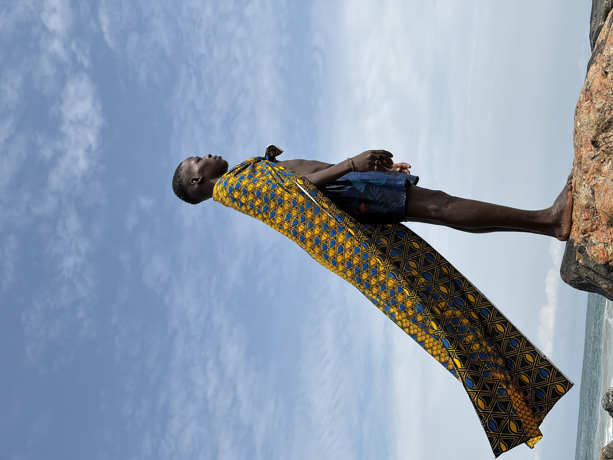 A young Black male stands on a rocky shore, his body angled to the right, facing the ocean. He is wearing blue patterned swim shorts and a vibrant, patterned fabric draped over his shoulders, creating a cape that extends dramatically behind him. The fabric, predominantly yellow and blue, is adorned with geometric and organic motifs. The sky above is a light blue, dotted with scattered white clouds. To the lower left, a sliver of the ocean and shoreline is visible. The man has short, dark hair and a focused expression. He is wearing a ring on his left ring finger and a thin bracelet on his left wrist. The scene appears to be an outdoor location, possibly a coastal area, consistent with the environment of Alaguntan, Nigeria.