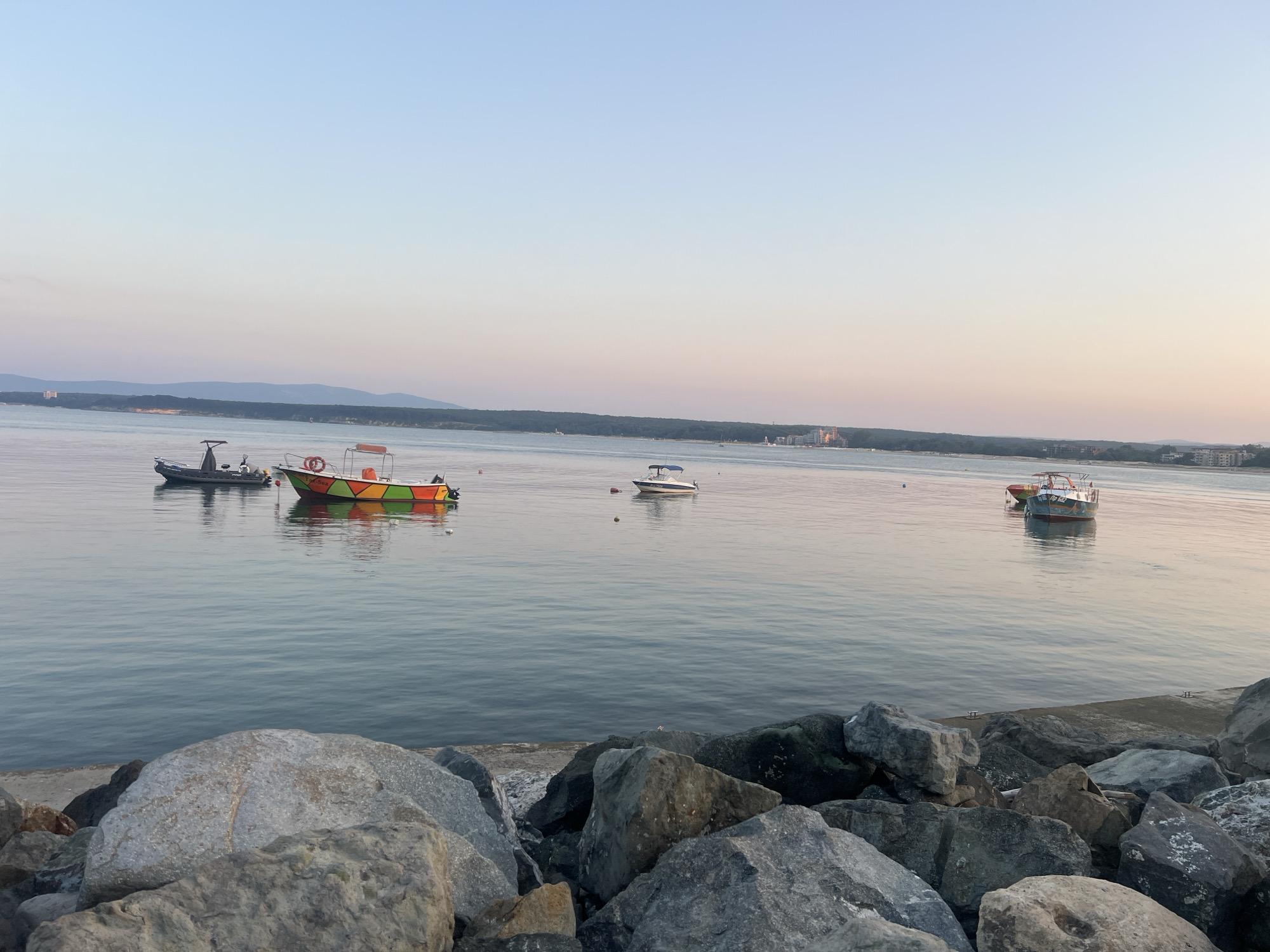 The image depicts a serene seascape with several boats anchored in the calm waters off the coast of Primorsko, Bulgaria. The sky is a gradient of soft blues and pinks, suggesting either dawn or dusk. The foreground is dominated by a rocky shoreline, with large, weathered stones of various shades of gray and brown creating a rugged texture.
In the middle ground, the sea stretches out, reflecting the pastel hues of the sky. Four boats are visible. The closest to the left is a dark-colored vessel with a distinctive upright fin. Next to it, a brightly colored boat, adorned with orange, green, and red geometric patterns, floats on the water, a lifebuoy visible on its deck. Further out, a smaller, white boat with a blue canopy is present. On the far right, another boat with a colorful hull, possibly red and blue, is anchored.
The background reveals a distant coastline characterized by a line of trees and scattered buildings, hinting at the presence of the town of Primorsko. The overall atmosphere is tranquil and picturesque, capturing a quiet moment by the Black Sea.