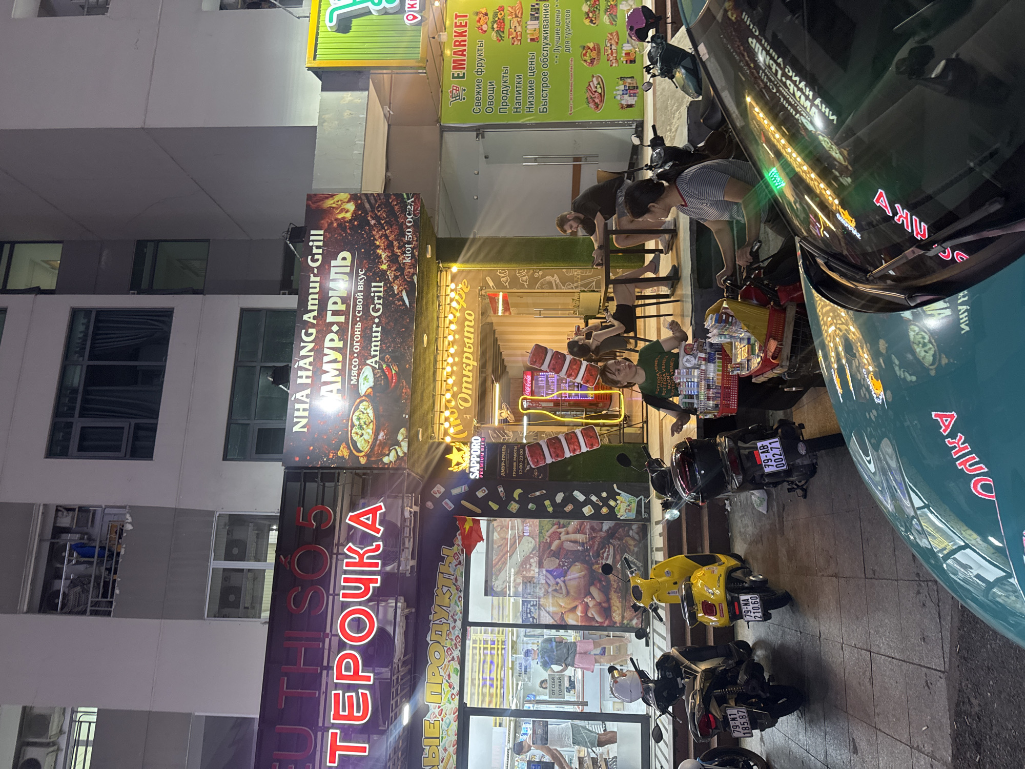 This is a vertical, nighttime street-level photograph taken in Bac Nha Trang Ward, Vietnam. The scene captures a bustling street with several shops and restaurants. The primary focus is on the storefronts adorned with neon signs and illuminated displays. One prominent sign reads "ТЕРОЧКА" in large red Cyrillic letters, and another advertises "NHÀ HÀNG Amur-Grill" with images of grilled food.
Several people are visible, mostly in the foreground and mid-ground, appearing to be customers or staff. Some are standing, others are seated at outdoor tables, and one person is tending to a motorcycle. The overall atmosphere suggests a lively commercial area in the evening. Various objects are present, including motorcycles parked along the sidewalk, shopping baskets, and shelves stocked with goods in the shop windows.
The time of day is clearly night, indicated by the artificial lighting and the dark sky. The weather appears clear, as there are no visible signs of rain. The location is suggested by the Vietnamese text on some signs ("NHÀ HÀNG" means "restaurant") and the style of the motorcycles, common in Southeast Asia. The mood seems active and social, with people engaged in dining or shopping activities. Text visible includes shop names, menu items, and promotional messages, some in Cyrillic and some in Vietnamese. For example, one sign says "EMARKET" and lists items like "Свежие фрукты" (fresh fruits) and "Продукты" (products). Another sign has "Coca-Cola" branding.