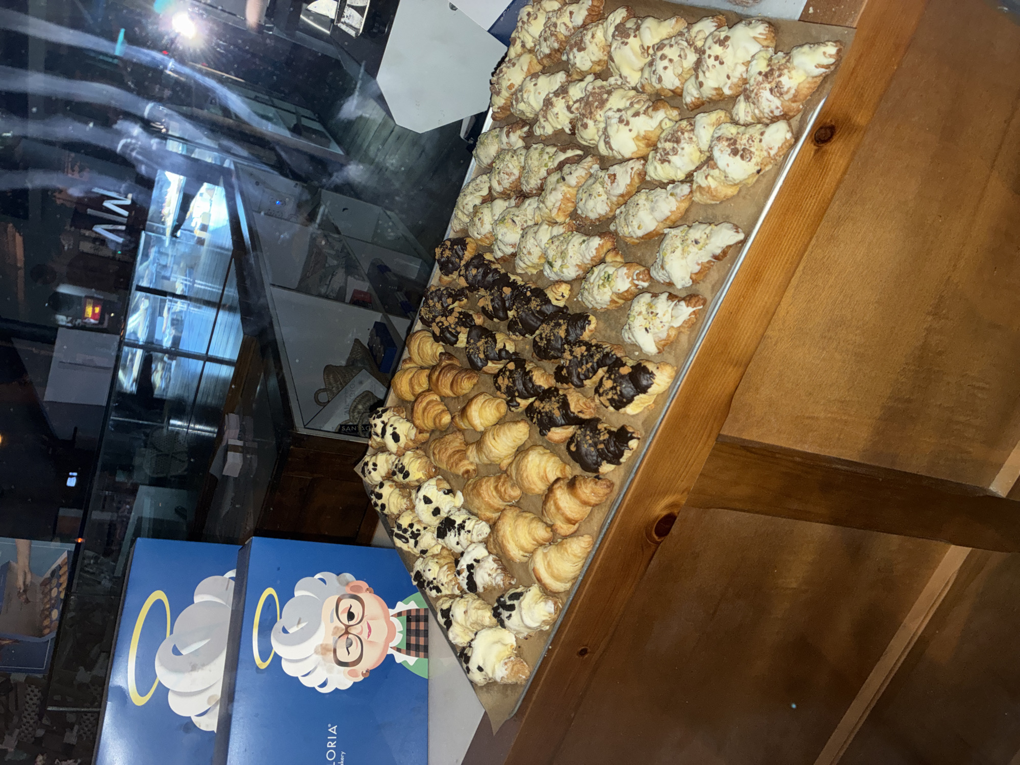 The image displays an interior scene, likely a bakery or cafe, specifically identified as La Gloria Bakery in Barcelona, Spain, based on visible branding. The foreground features a light brown wooden countertop. On the counter, a large rectangular metal baking sheet, covered with brown parchment paper, holds numerous mini-croissants arranged in rows. These pastries exhibit various toppings: some are plain golden-brown, others are partially dipped in dark chocolate with crushed brown toppings, and a significant portion is covered in white frosting or glaze. The white-frosted croissants feature different garnishes including green crushed toppings (likely pistachio), brown sprinkles, and dark chocolate chips.
In the mid-left background, two blue cardboard boxes are stacked. The lower box prominently features a cartoon illustration of an elderly woman with white, cloud-like hair, a yellow halo, glasses, and a green plaid apron. The partial brand name "LORIA" is visible, identifying it as La Gloria Bakery. The top box displays only the cloud and halo design. The background on the right is dominated by a large glass pane, reflecting the illuminated interior of the establishment, including display cases holding pastries, shelves, and a bright light source causing glare. A reflected white sign with the characters "Λ/N" or "AW" is also visible. No individuals are directly visible in the immediate scene.
