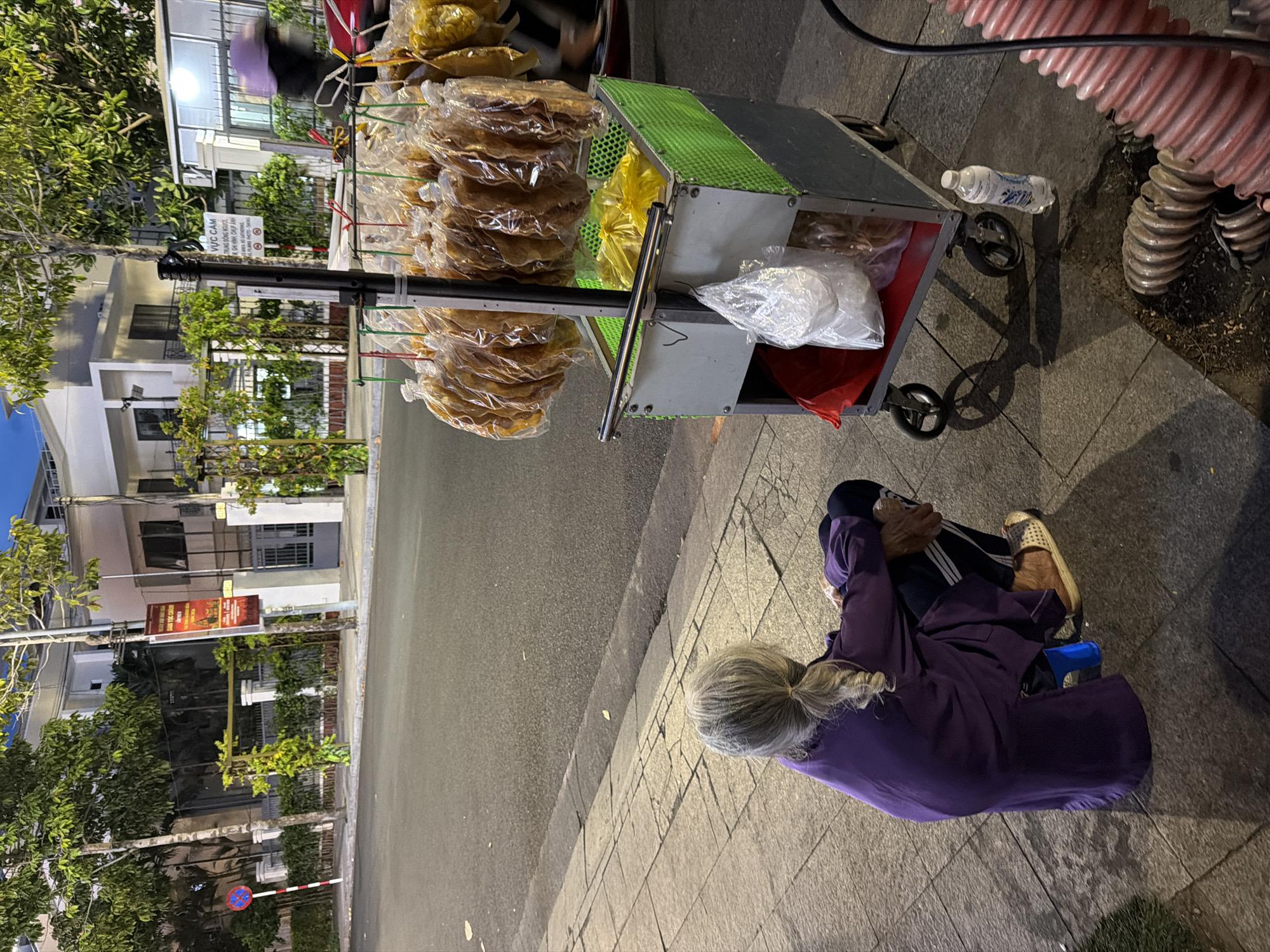In Nha Trang Ward, Vietnam, an elderly person with gray hair braided in a ponytail is sitting on a sidewalk next to a street. They are wearing a purple robe and dark pants, with a blue stool beneath them. To their right is a food cart displaying numerous bags of dried goods, likely snacks, hanging from a rack. The cart itself is metallic with a green mesh basket on top, holding yellow plastic bags filled with items. A plastic water bottle is on the ground next to the cart. In the background, modern buildings with white facades and trees line the street. The scene suggests a street vendor operating in the evening, with artificial lighting illuminating the area.
