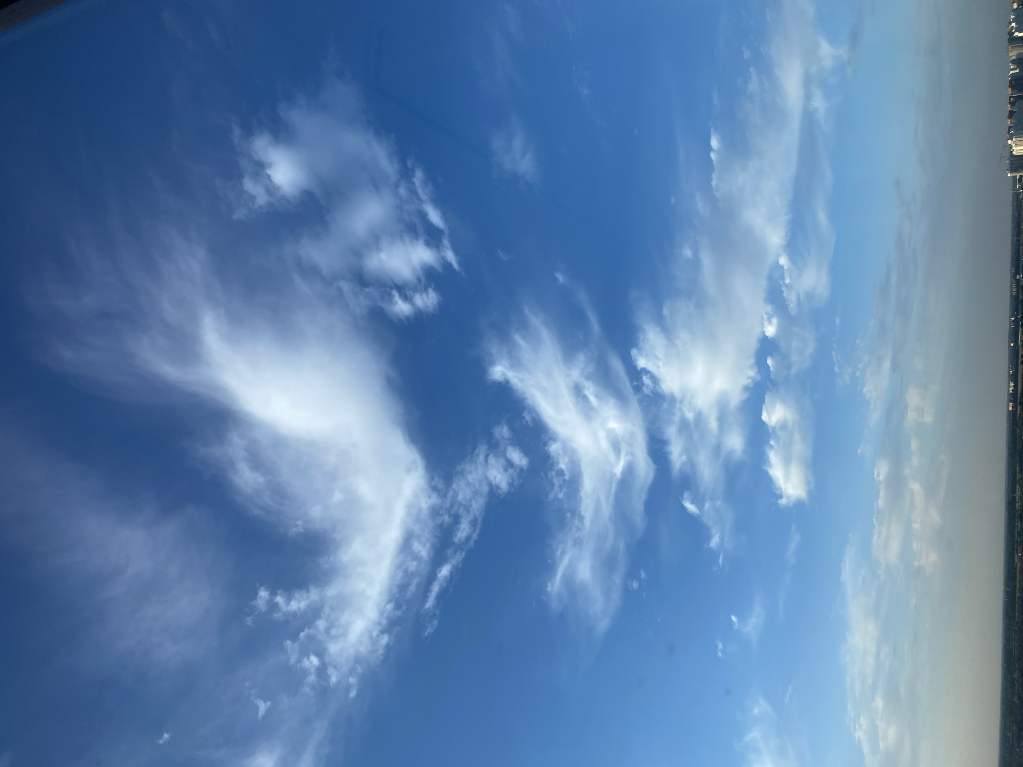 The image shows a wide view of a blue sky filled with scattered white clouds. Some of the clouds are wispy and thin, while others are more dense and fluffy. Towards the bottom of the image, a hazy horizon can be seen, suggesting the presence of a city or landscape in the distance. The lighting indicates it is likely daytime, possibly late afternoon or early morning given the haze. There are no people or animals visible in the frame. The overall impression is one of a vast, open sky above a distant urban or natural setting.