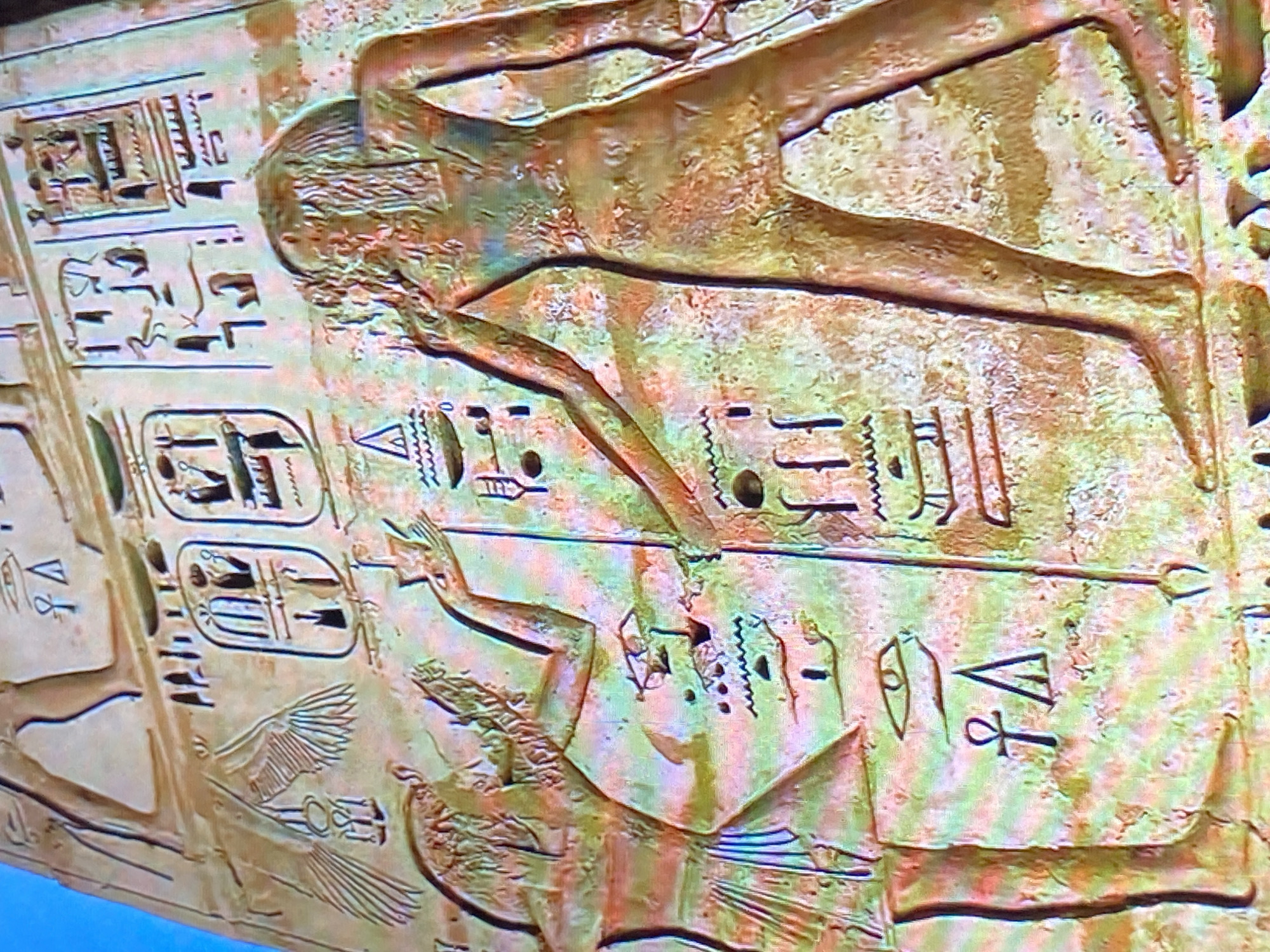 The image shows a section of an ancient Egyptian relief carving. The carving is on a stone surface, likely part of a wall or sarcophagus. Two figures are depicted in profile. The upper figure is elongated and appears to be horizontal. The lower figure is also elongated and horizontal, and has what seems to be wings. There are hieroglyphs alongside and between the figures. The background is colored with varying shades of brown and green. The photograph appears to be an unedited snapshot of a television screen displaying an image of Egyptian art. The city of Tinaquillo, Venezuela is not depicted.