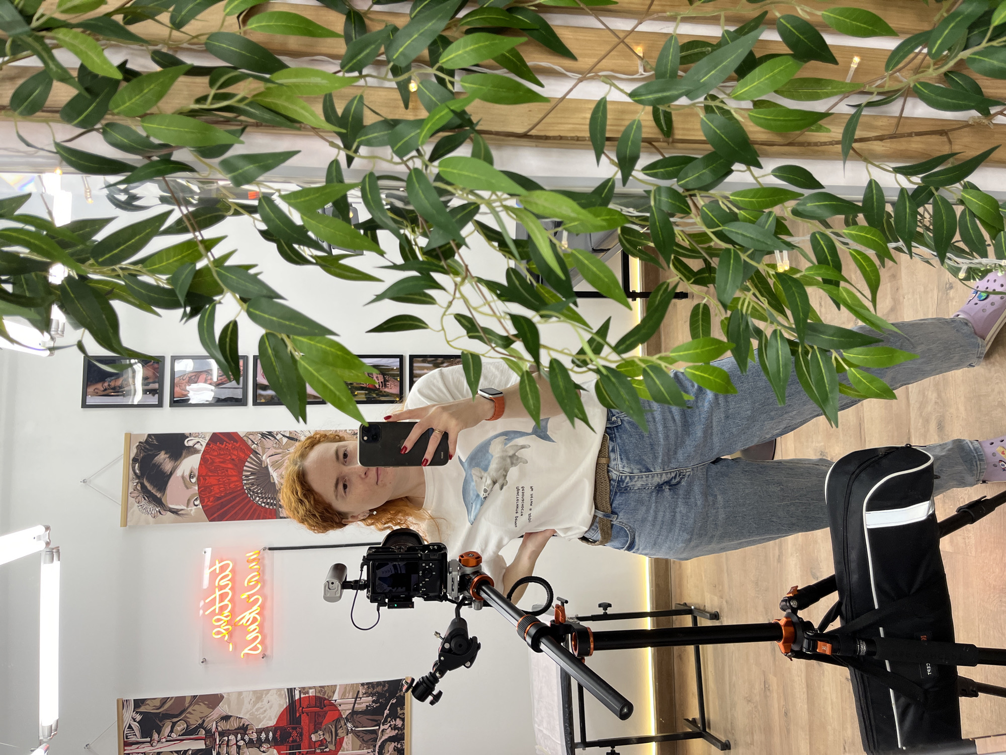 A woman with red hair is taking a selfie in a mirror. She is holding a black smartphone and wearing a white t-shirt, blue jeans, and purple shoes. In front of her is a camera on a tripod. Behind her, the wall is decorated with pictures, posters, and a neon sign in Russian. Faux greenery with fairy lights decorates the top of the image. The location is Chaikovsky, Russia. The floor is wooden.