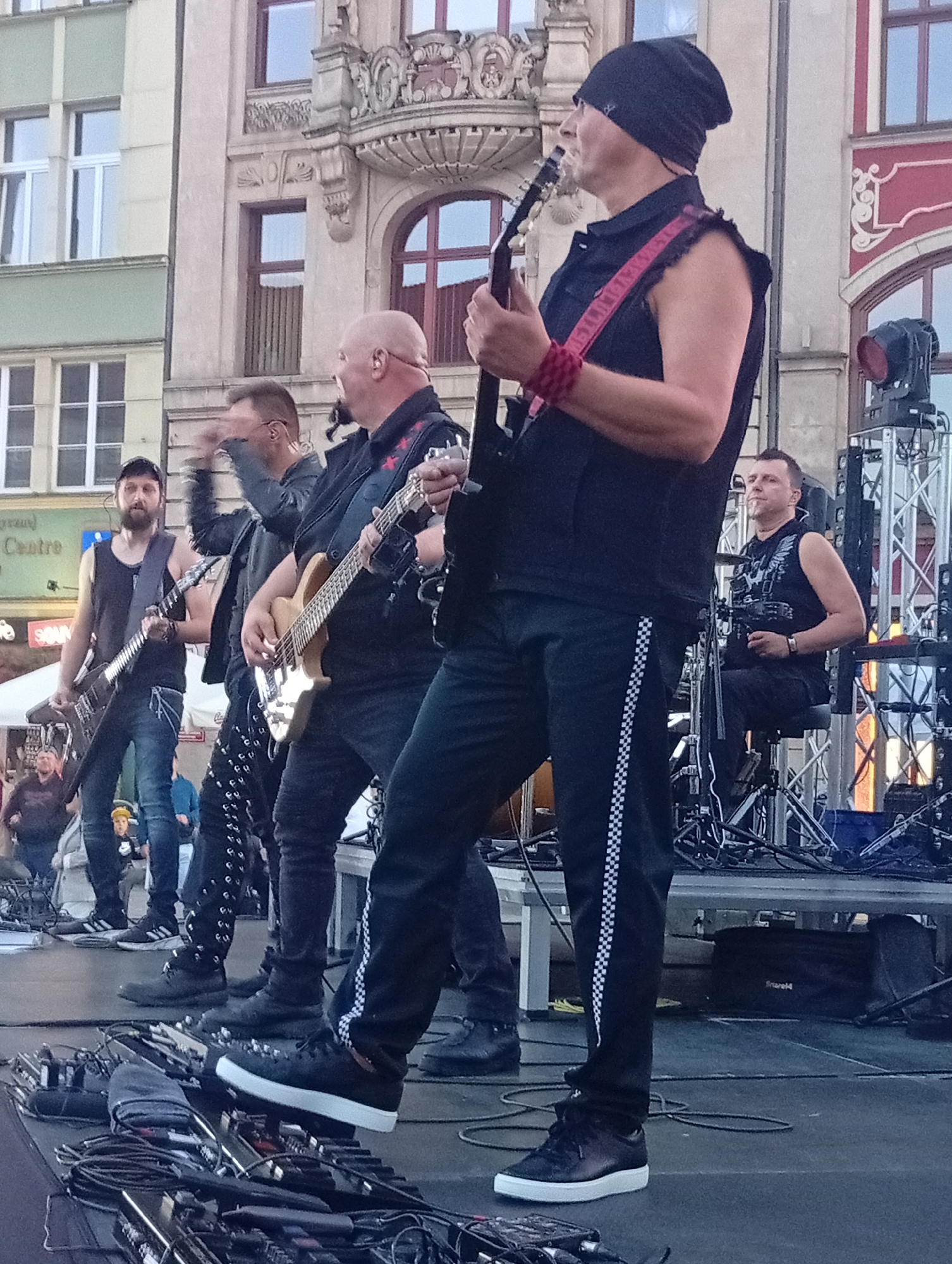 A group of male musicians are performing on a stage in front of an ornate building, likely in Wrocław, Poland. The building has multiple windows, some of which appear to be arched, and decorative architectural elements. The stage is set up with musical equipment, including guitars, amplifiers, and what appear to be pedalboards and cables on the floor in the foreground.
The musicians are dressed in dark, possibly black, clothing. One musician in the foreground, wearing a black beanie, is playing a guitar and has a red wristband. Another musician to his left is also playing a guitar. Further back, a bald musician is playing a bass guitar. Another musician, partially obscured, is playing drums. The atmosphere appears to be an outdoor concert or festival. The lighting suggests it might be late afternoon or early evening.