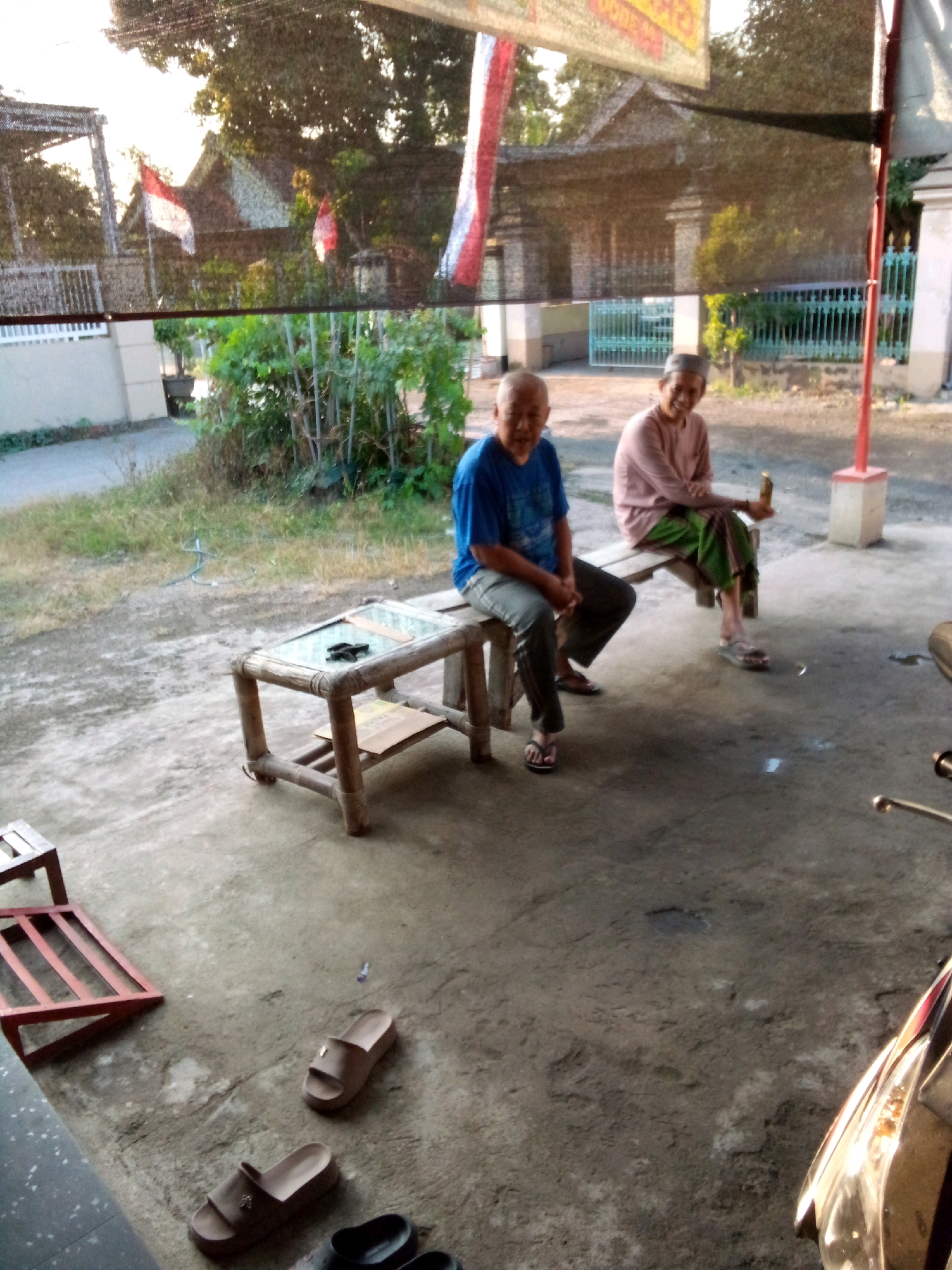 Two elderly individuals are seated outdoors. One man, wearing a blue shirt and gray pants, sits on a wooden stool next to a small bamboo coffee table. The other person, wearing a pink shirt and green pants, sits on a wooden bench. They are in what appears to be a residential area in Simo, Indonesia, with a building and vegetation visible in the background. The ground is concrete and there are several pairs of sandals scattered around.