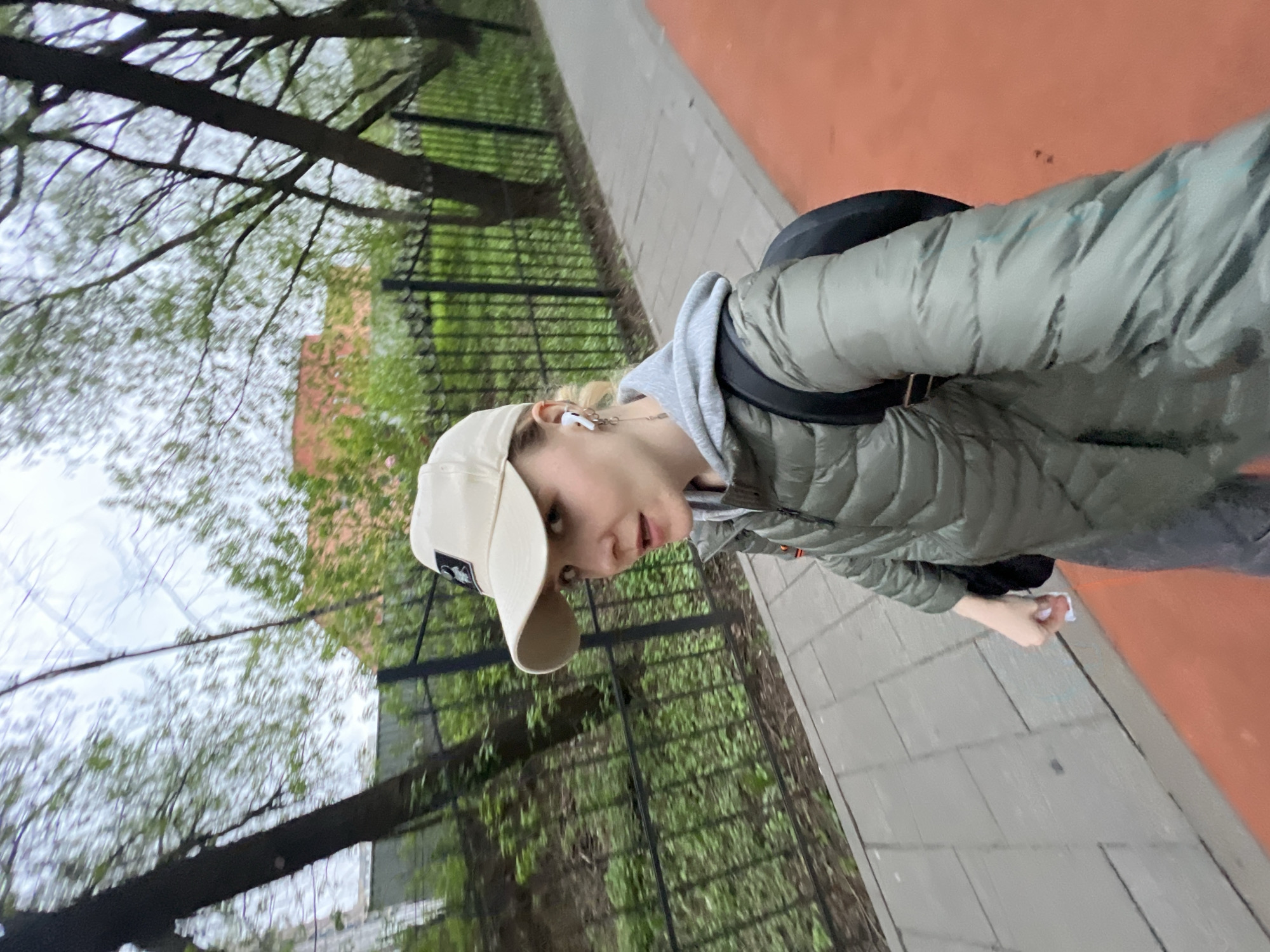 A woman stands on a gray tiled walkway adjacent to a red paved area. A black metal fence runs parallel to the walkway. Behind the fence, there is green vegetation and a glimpse of a red brick building. Bare trees with budding leaves are overhead. The woman is wearing a beige baseball cap, a gray hooded sweatshirt, and a green puffer jacket, with a black backpack. She is looking at the camera. The location is Khimki, Russia.