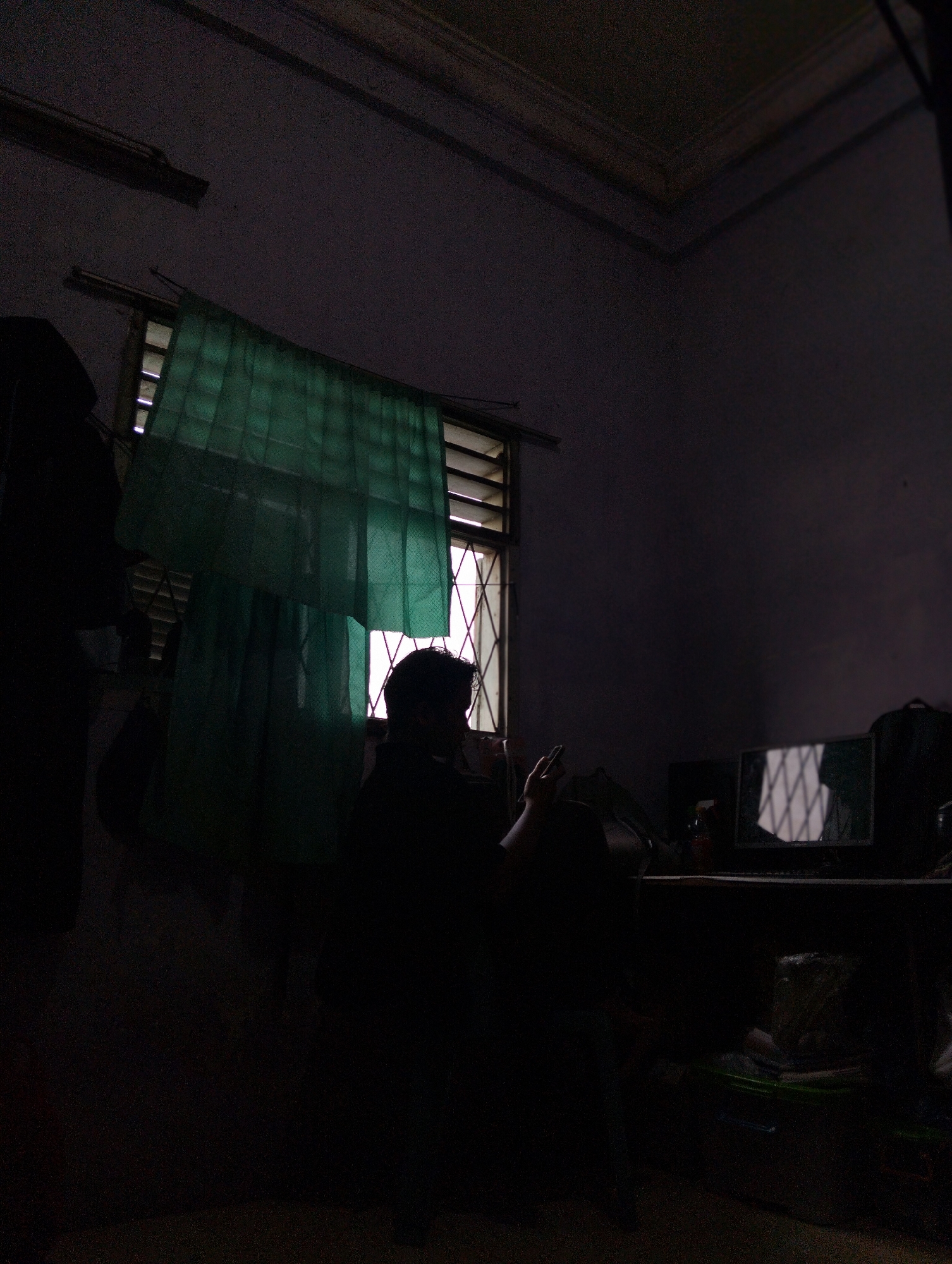 An over-the-shoulder, medium shot captures a person silhouetted against a window in a dimly lit room. The individual, facing away from the camera, is seated at a desk and appears to be holding a mobile device. To the left, a green curtain hangs across the window, filtering the natural light. On the desk, a computer monitor displays a reflection of the window's diamond pattern. The room's interior is dark, with the ceiling showing ornate trim. The scene suggests a domestic or personal space, possibly a bedroom or study, in a building located in the City of Medan, Indonesia.