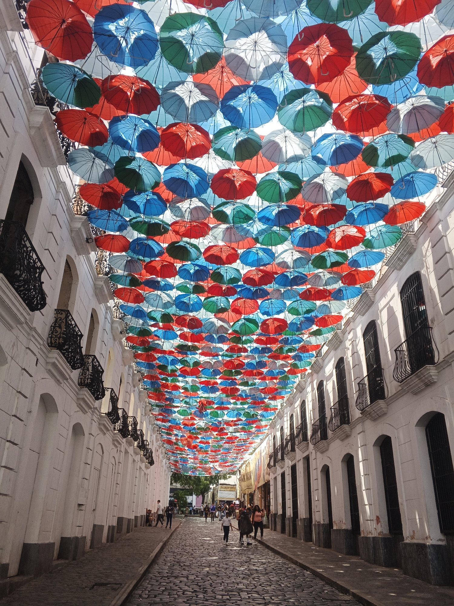 A long, narrow streetscape in Caracas, Venezuela, is covered by a canopy of hundreds of colorful umbrellas - red, blue, green, and white - suspended overhead. The street, constructed of rough-hewn paving stones, is lined on both sides by white buildings with arched windows and ornate black iron balconies. Several people are walking along the street, with some individuals and groups clustered together. The time of day appears to be daytime, with dappled sunlight filtering through the umbrella canopy onto the street and buildings. The overall scene is vibrant and lively, emphasizing the architectural surroundings and the colorful umbrella installation.