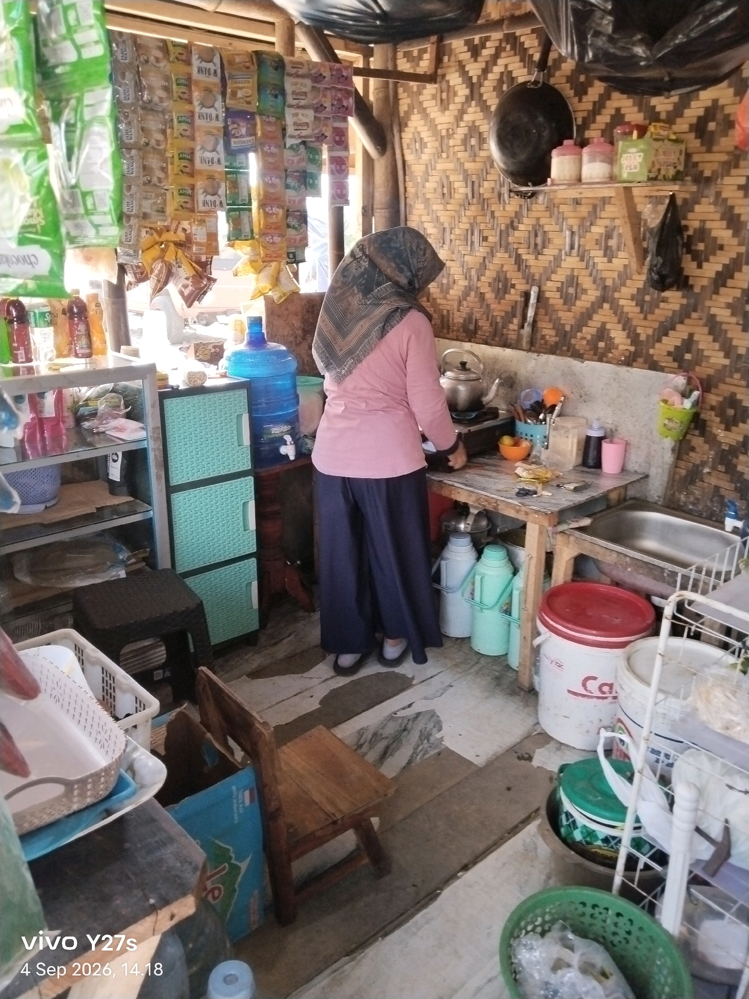 In the city of Sigedang, Indonesia, a woman wearing a headscarf and a pink long-sleeved shirt stands in a rustic, open-air kitchen, possibly a warung or small eatery. The wall behind her is constructed from woven bamboo. She is positioned in front of a small stovetop where a kettle is boiling, suggesting she is in the process of preparing a hot beverage or cooking.
The kitchen is compact and cluttered with various items. Shelves on the left are laden with packaged goods, likely snacks or instant noodles, and several bottles of beverages. To the right of the stovetop is a small wooden table with a sink. On the table, there are bowls, utensils, and containers, including a blue plastic basket filled with items. Below the table, several large plastic water dispensers and buckets are visible, including a red-lidded white bucket.
In the foreground, to the left, a white plastic basket with a lattice design sits on a wooden surface. To the right of this, a small, simple wooden stool is placed near a large blue cardboard box. The floor appears to be made of concrete tiles. The overall environment suggests a functional, somewhat makeshift kitchen area where food and drinks are prepared for sale or consumption. The lighting indicates it is daytime, with natural light entering from the left.