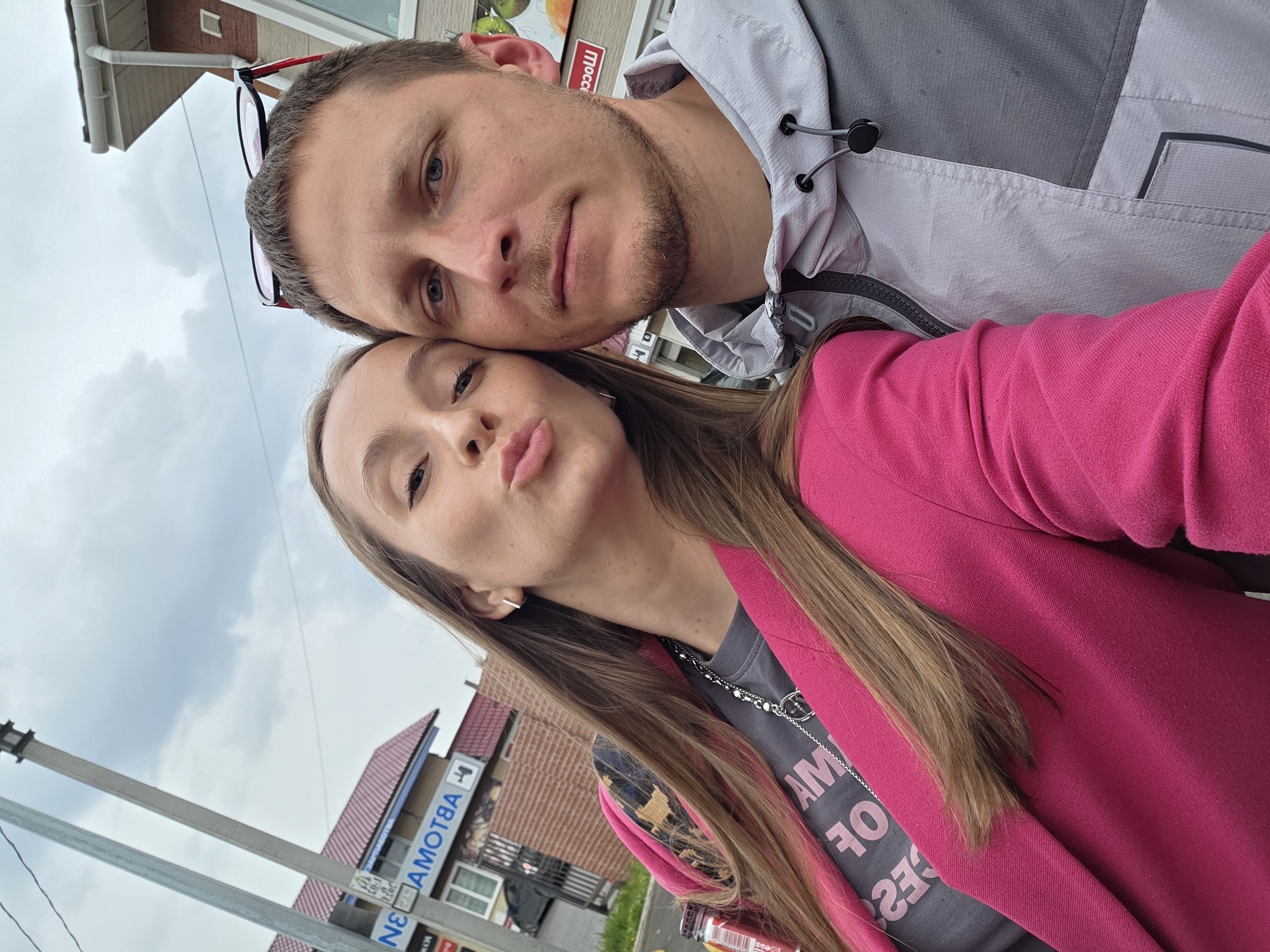 A man and a woman are posing for a selfie outdoors in what appears to be Nikolayevka, Russia. The man has short brown hair, light skin, and is wearing a gray hooded jacket. He has glasses pushed up on his forehead. The woman has long brown hair and is wearing a pink blazer over a gray t-shirt with white lettering. She is making a kissing face toward the camera.
In the background, there is a building with a red tile roof and a sign in Russian that appears to say "AVTOMOTIV". There is also a utility pole and a cloudy sky visible. A beverage can sits at the bottom of the frame.