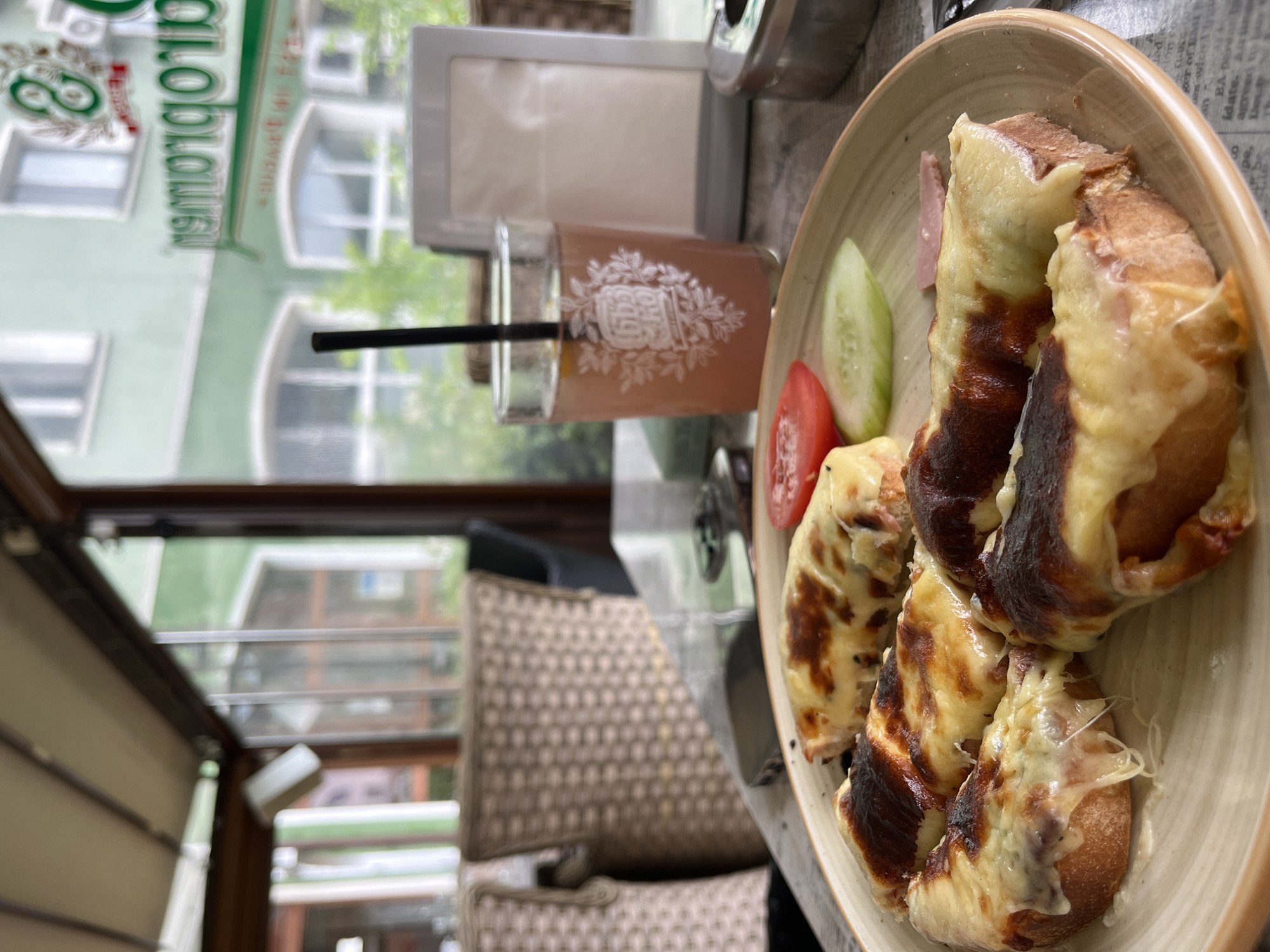 The image depicts a close-up view of a plate of open-faced sandwiches topped with melted cheese and some charred bread. The sandwiches are arranged on a light-colored plate with a subtle pattern. Accompanying the sandwiches are a slice of tomato and a slice of cucumber. A thin slice of ham is also visible on the plate. In the background, a glass of pinkish liquid with a black straw is placed on a table. To the right of the glass, a white, rectangular object resembling a photo frame or a placeholder is visible. An ashtray with a partially obscured contents is situated to the far right. The scene appears to be set on a table covered with a newspaper. Beyond the table, a blurred background suggests an outdoor seating area of a cafe or restaurant, with wicker chairs and a view of buildings through a glass enclosure. The architectural style of the buildings and the signage in the background suggest a European setting. Specifically, the building with green facade and the visible sign written in Cyrillic script points to a location in Bulgaria. The sign reads "RODOPOLOV", which is a surname common in the Rhodope Mountains region of Bulgaria. Considering the typical offerings of cafes in such regions, this establishment is likely located in the city of Rudozem, Bulgaria, known for its proximity to the Rhodope Mountains.