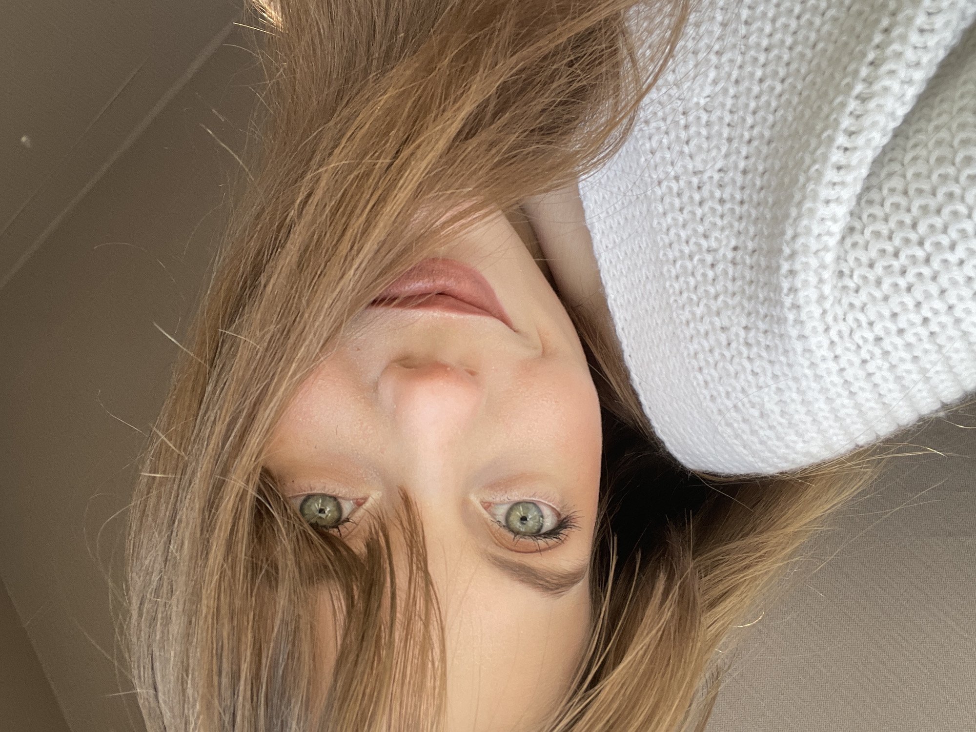A close-up, upside-down portrait of a young woman with light brown hair. Strands of hair partially obscure her face, particularly around her left eye (from the viewer's perspective). Her eyes are green. She has a light complexion and light pink lips. She is wearing a white, knitted sweater. The background is a neutral beige tone. There is no discernible information to indicate the location is Ufa, Russia.