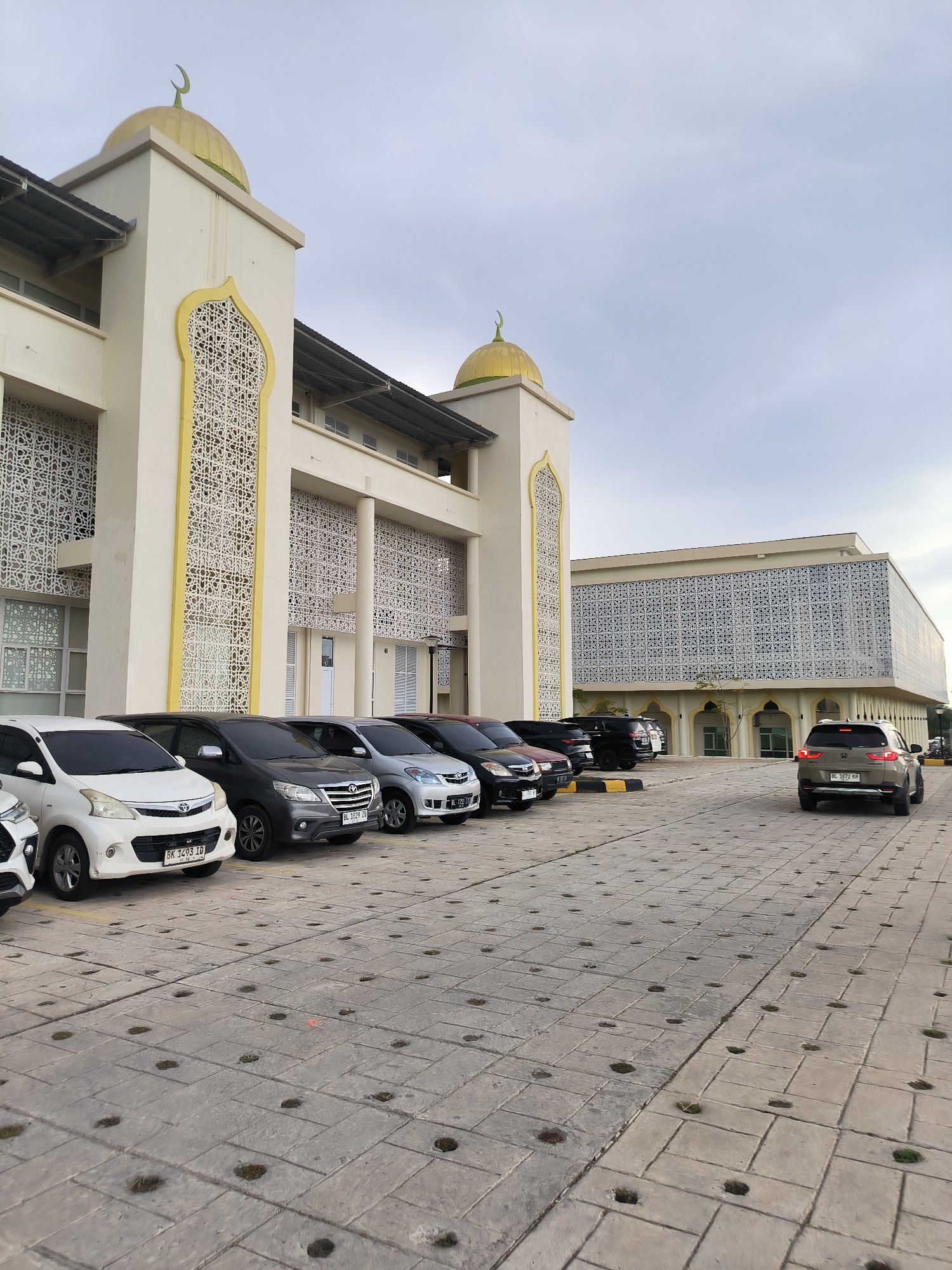 The image depicts the exterior of a mosque in Reuleut Timu, Indonesia. The mosque features a cream-colored facade with prominent golden domes topped with crescent moons. Intricate white latticework adorns the building's exterior, particularly around arched openings. In front of the mosque, a parking lot is visible, occupied by several cars parked in a row. One car, a grey SUV, is shown driving away from the viewer on the right side of the frame. The ground in the foreground is paved with textured concrete tiles, with small circular openings scattered across the surface, some containing patches of grass. The sky is overcast, suggesting a cloudy day.