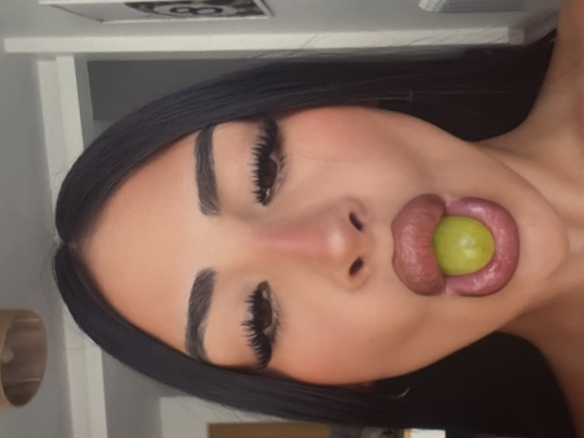 A close-up, waist-up shot shows a woman's face with dark hair. She is wearing makeup and has a green grape in her mouth. Her lips are full and appear to be tinted. The background is blurred and shows a white wall with a framed picture on it. The lighting is soft and diffused. The image is likely taken indoors.