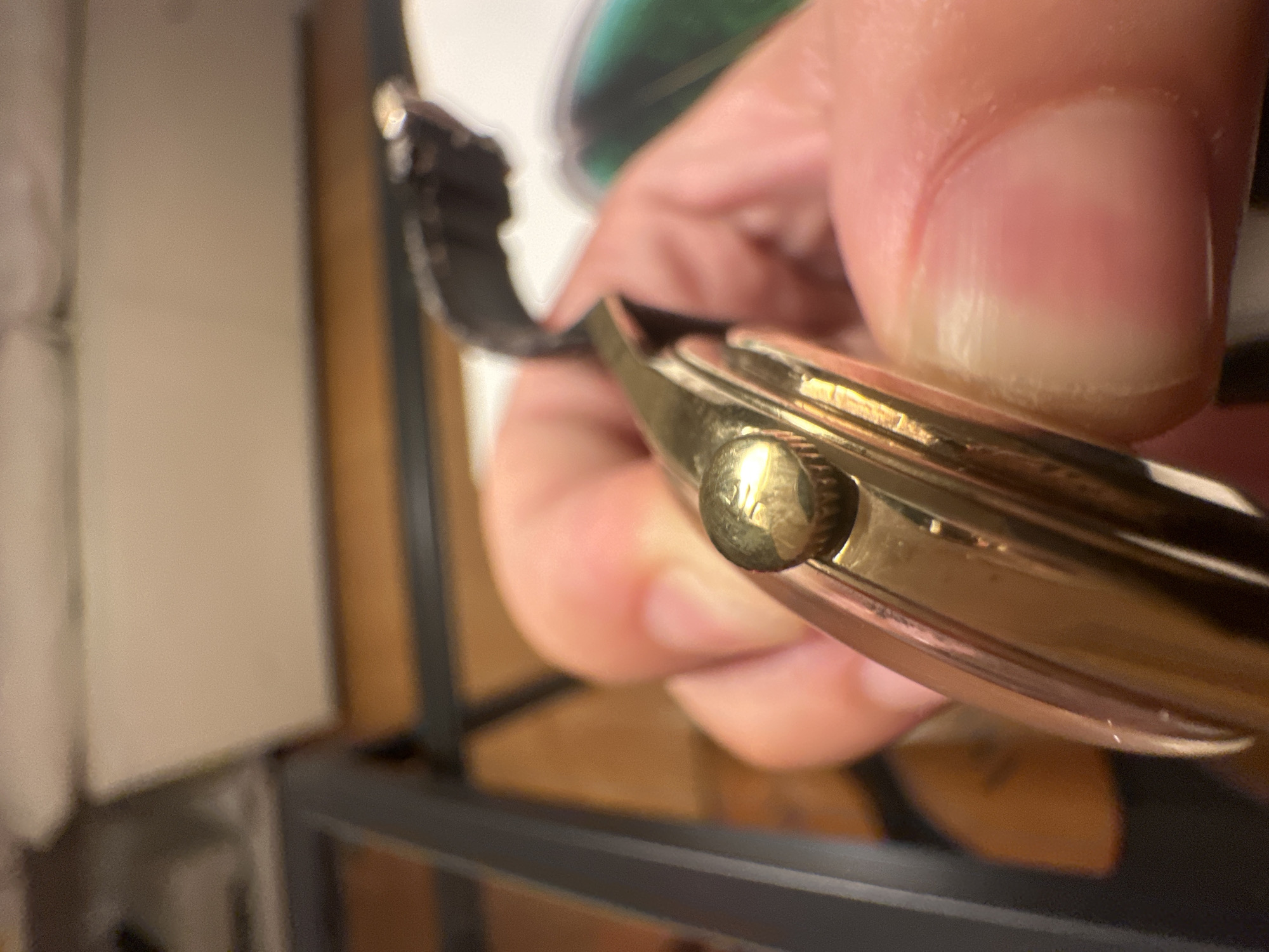 The image displays a close-up perspective of a human hand holding a wristwatch. The hand, exhibiting light-toned skin, grips the watch from the side, with the thumb and forefinger prominently visible. The wristwatch features a gold-toned case with visible signs of minor wear and superficial scratches. A prominent, gold-toned fluted crown with an etched symbol (indistinguishable due to resolution but appears to be a stylized 'T' or similar character) is located on the right side of the watch case, positioned between the fingers. A dark-colored watch strap extends from the case into the background. Slight redness is visible on the distal phalanx of the thumb, indicative of pressure. The background is entirely out of focus, showing blurred shapes and colors consistent with an indoor environment. Distinguishable background elements include blurred light-colored surfaces, darker linear structures, and a green-tinged area in the far right. No discernible details within the image allow for the identification of a specific location within the city of Milan, Italy.