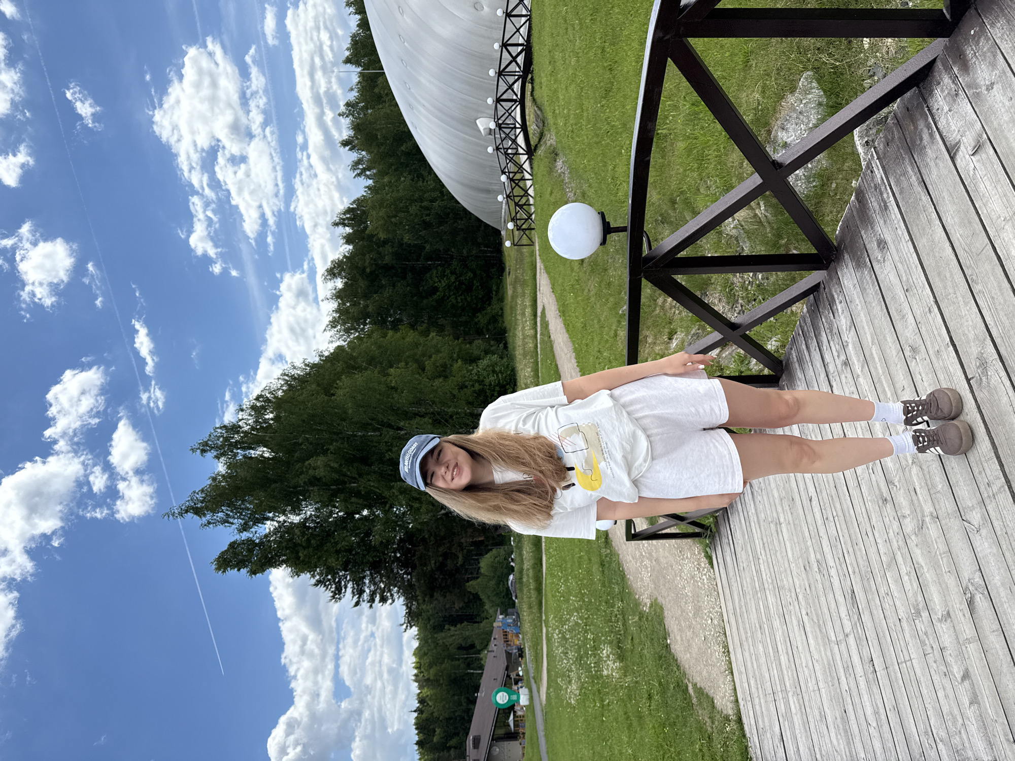A young woman with long blonde hair and a cheerful smile is standing on a wooden deck, slightly leaning against a black metal railing. She is wearing a casual outfit consisting of a white t-shirt with an abstract graphic design (featuring a prominent yellow shape), white shorts, white socks, and brown sneakers with white accents. A blue baseball cap, with partial white text "ATLANTIC OCEAN" visible, sits on her head.
The scene is outdoors, likely a recreational or park area during daytime. Behind the woman, to her right, a large white dome-shaped building is prominent, set against a backdrop of green grass and dense green trees. A paved pathway runs alongside the grass, leading towards the dome. A white spherical lamp is mounted on the black railing. Further in the background, more trees and hints of other structures can be seen, including a building with a brown roof and a green circular sign.
The sky is bright blue with scattered white cumulus clouds. A distinct white contrail from an airplane stretches across the upper part of the sky. The lighting suggests a sunny day. The overall atmosphere is relaxed and pleasant. The location context indicates this scene is in Dobrograd, Russia.
