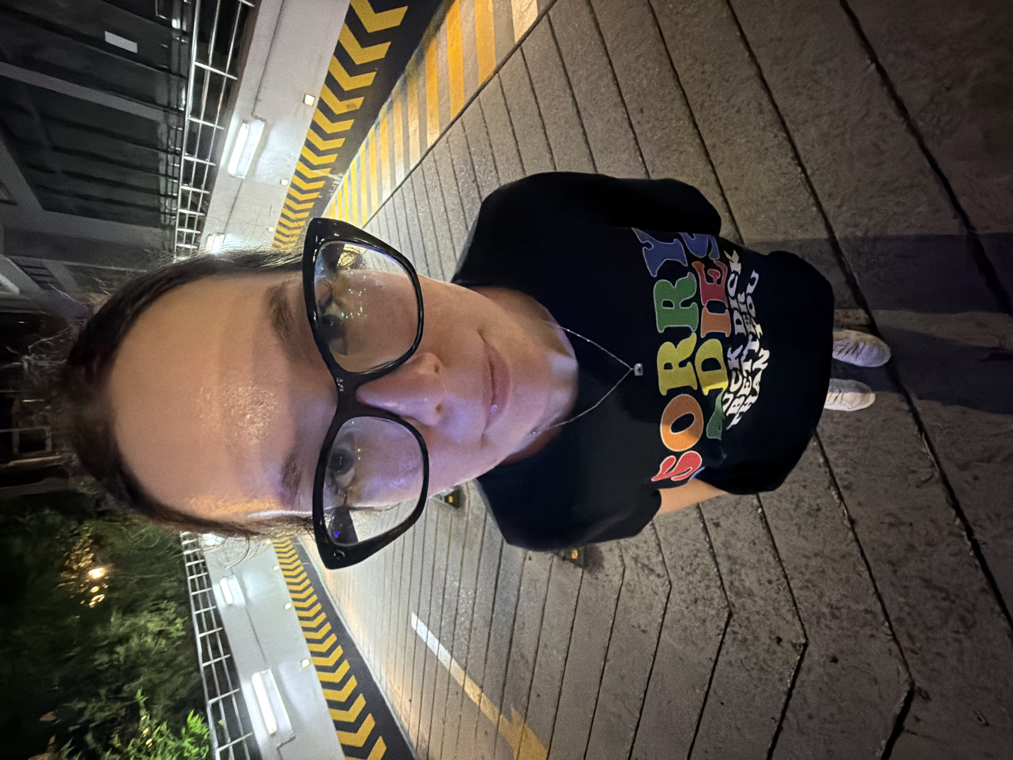 A medium shot, fisheye lens photograph, taken at night, shows a person wearing glasses and a black t-shirt with colorful lettering. The person is standing on a concrete ramp with yellow and black striped markings on the sides. The ramp appears to be an entrance or exit to an underground parking structure, as indicated by the overhead structure visible in the background. The overall impression is of an urban setting, likely in a city like Hanoi, Vietnam, given the presence of lush greenery and modern architecture in the background. The lighting suggests the photograph was taken under artificial streetlights.