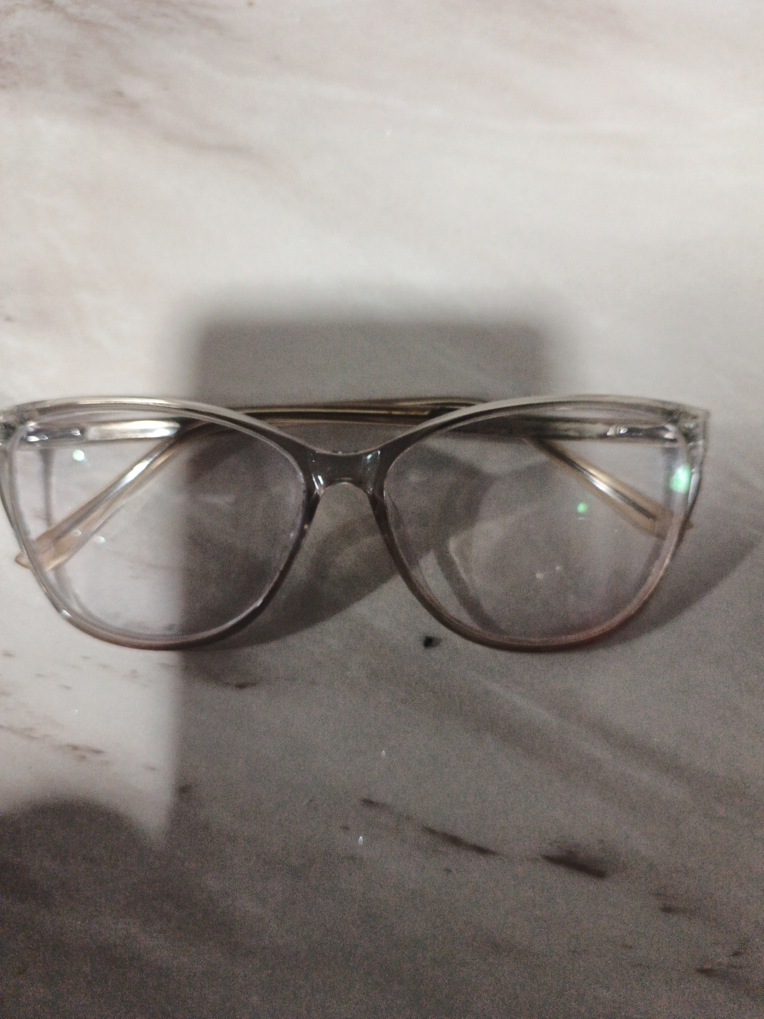 A pair of clear-lensed eyeglasses with thin, clear frames is centrally positioned on a speckled, greyish-brown surface. The eyeglasses are oriented horizontally, with the lenses facing the viewer. The surface appears to be a countertop or table, with subtle variations in color and texture, suggesting a marble or granite material. The lighting in the scene creates a shadow to the left and bottom of the eyeglasses, indicating the direction of the light source. There are no people visible in the image. The background is out of focus and appears to be a continuation of the same speckled surface. The image was taken in Kelapapati, Indonesia.