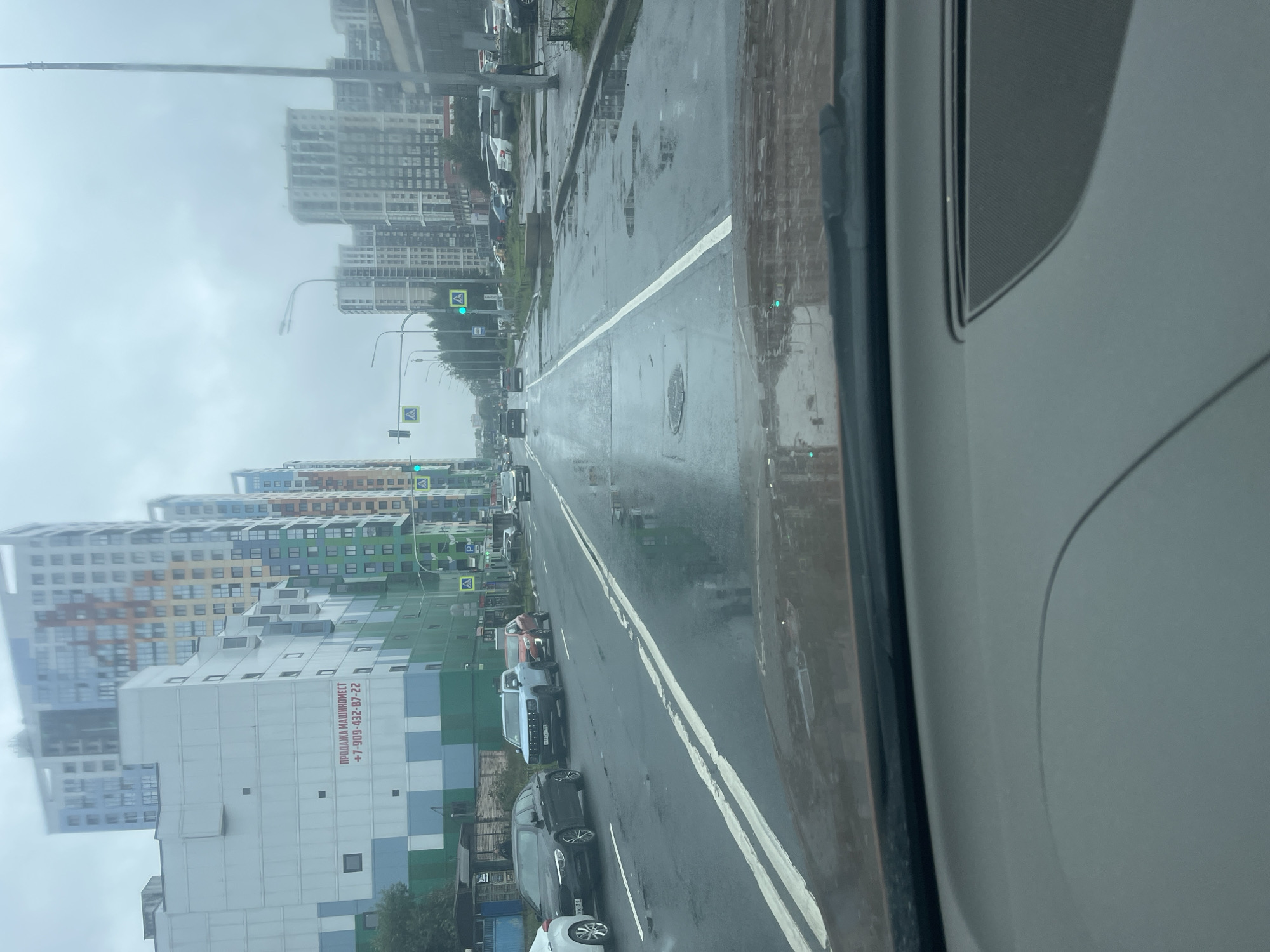 The image shows a view from inside a car, looking out at a wet street in Saint Petersburg, Russia, with several modern buildings in the background. The street is lined with cars, and the road surface is reflective due to the rain. Pedestrian crossings and traffic lights are visible. A large, multi-story building with a facade featuring a grid pattern of white, blue, and green squares is prominently displayed on the left side of the image, with Russian text and a phone number displayed on it. In the distance, a series of tall, colorful apartment buildings are visible. The sky is overcast and appears to be a hazy gray. The interior of the car, including part of the dashboard and windshield, is visible in the foreground. The overall scene suggests an urban environment during or after a period of rain.